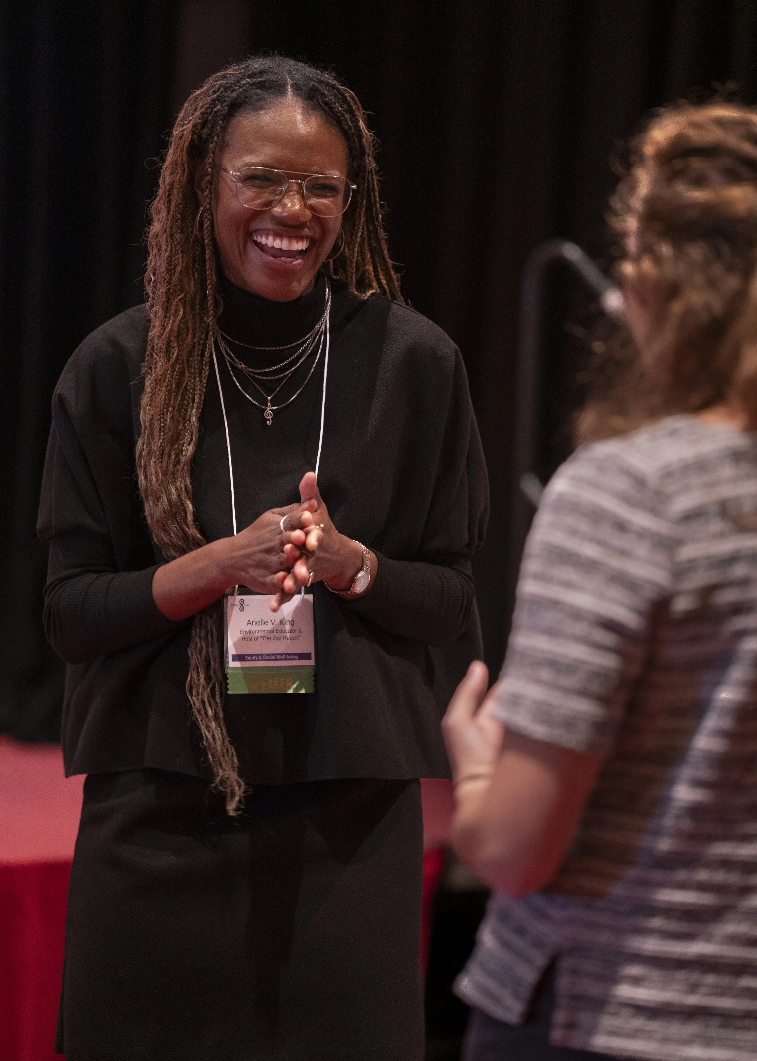 A woman with long dreadlocks and glasses, smiling and wearing multiple necklaces, talking to another woman with curly hair in a striped shirt at an indoor event.