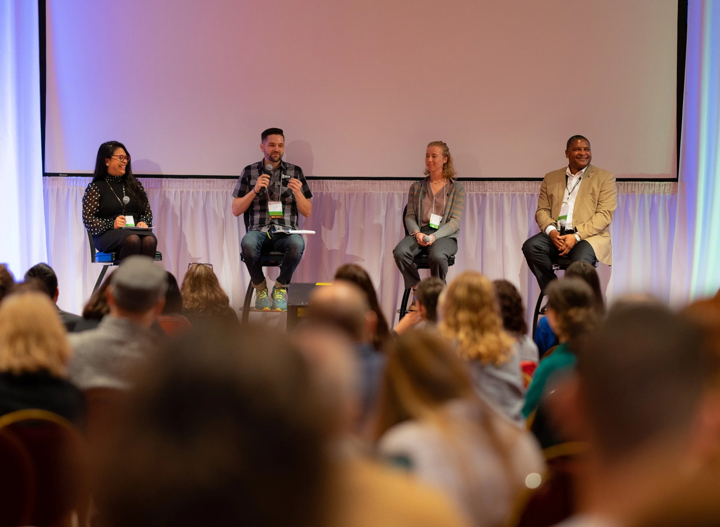 A panel of four speakers on stage at a conference, seated on stools, with an audience in front of them.