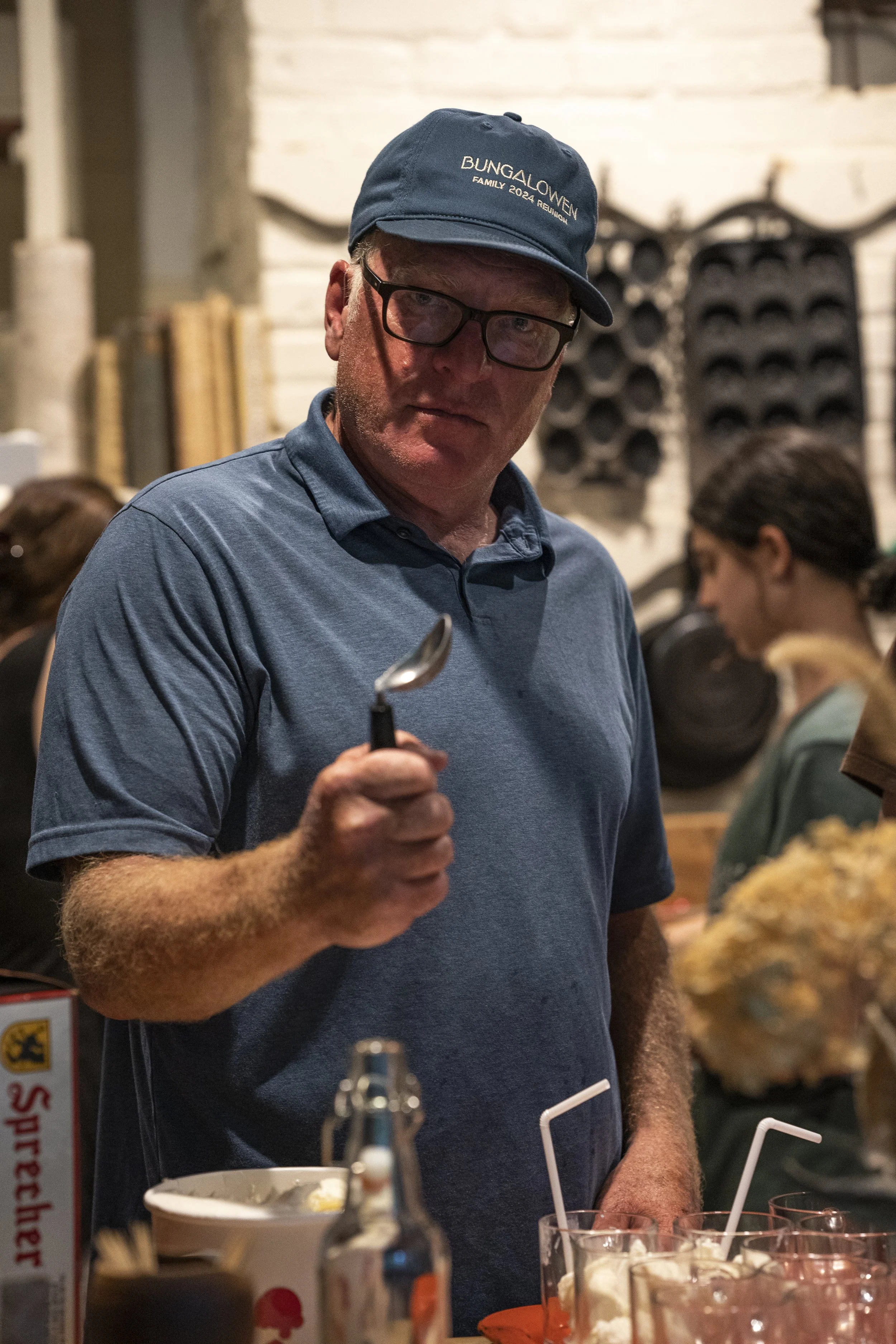 A man wearing glasses, a blue cap reading 'Bungalow 22 Family 2024 Reunion,' and a blue polo shirt, holding a spoon in a busy restaurant or bar setting.