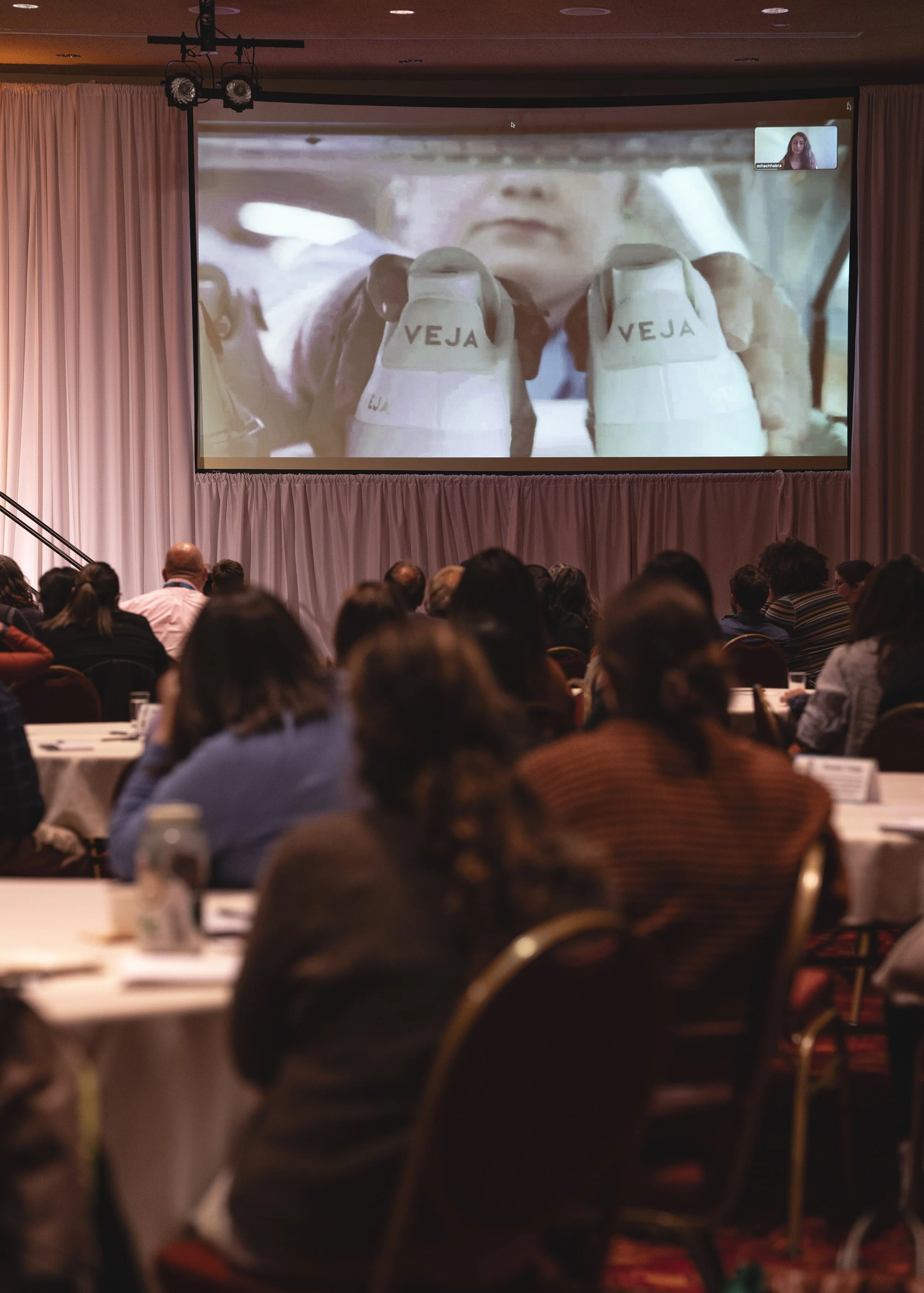 A conference room filled with attendees watching a presentation on a large screen. The screen displays an image of a person holding up two shoes labeled 'VEJA'. 