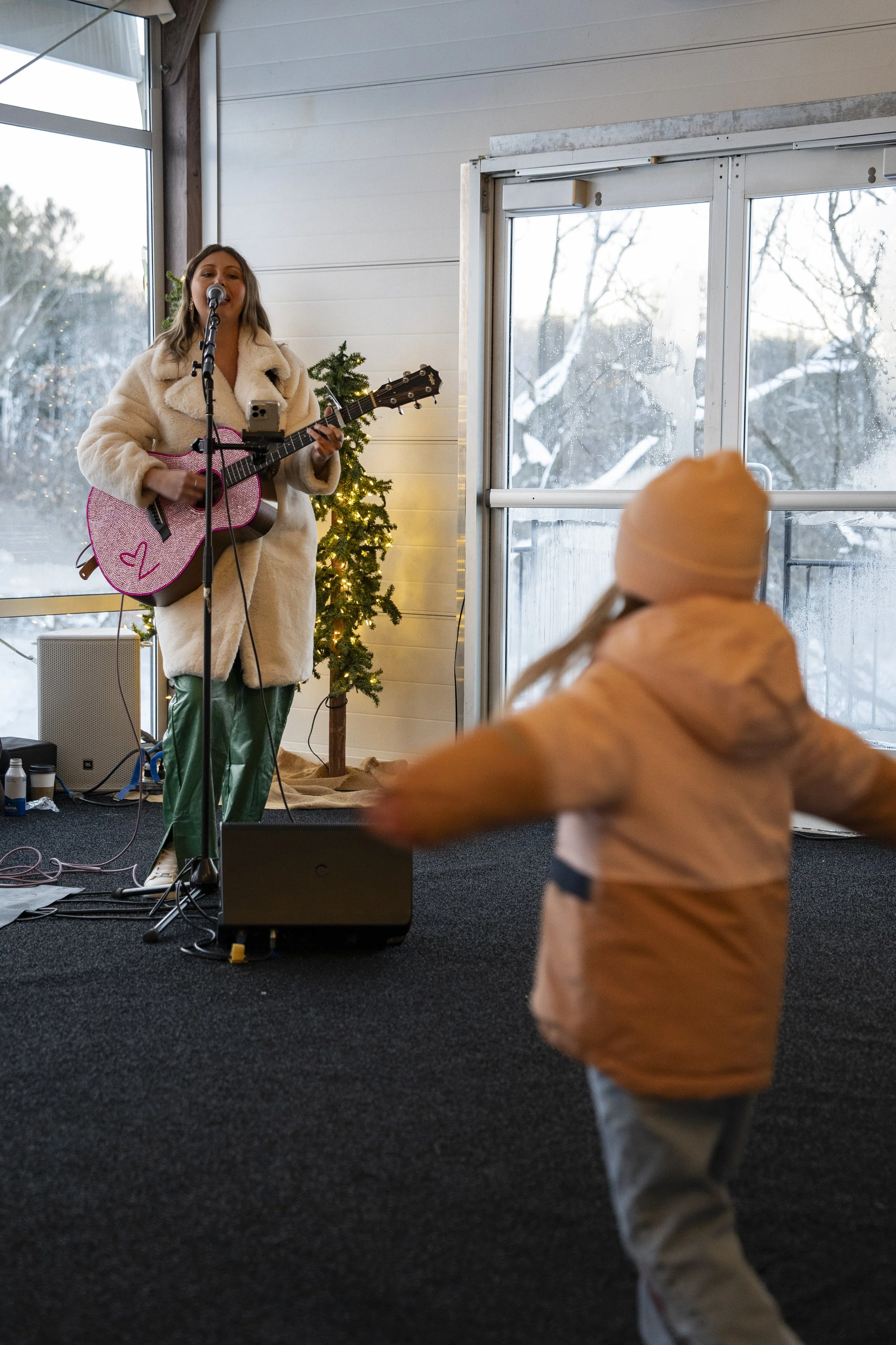 A woman singing and playing guitar on stage decorated with a small Christmas tree, while a child in a pink jacket and beige hat dances in front of her during a winter event.