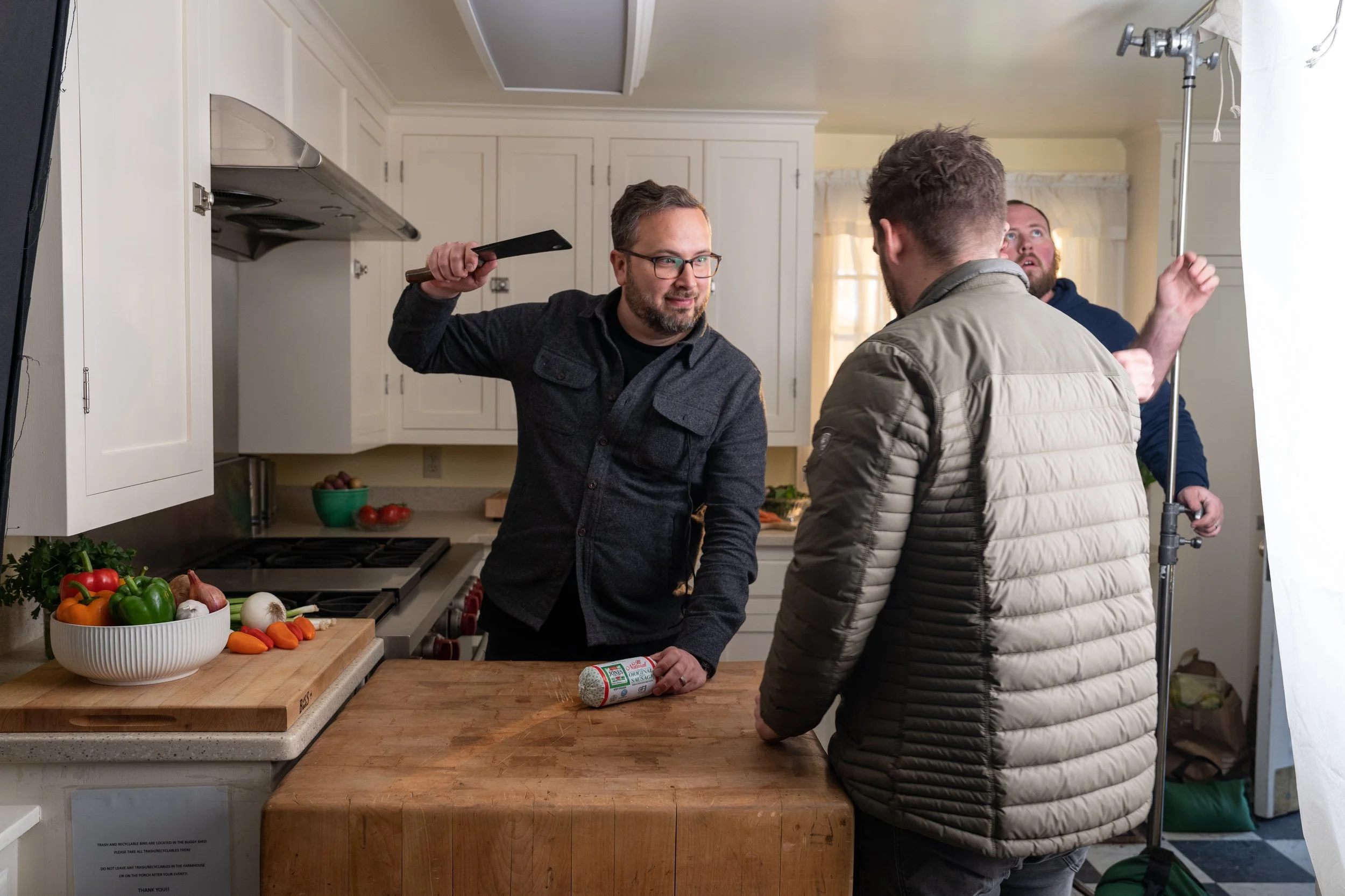 A behind-the-scenes view of a kitchen scene being filmed, featuring a man with glasses holding a kitchen knife and a sausage, facing another man wearing a beige jacket, with a crew member operating lighting equipment in the background.
