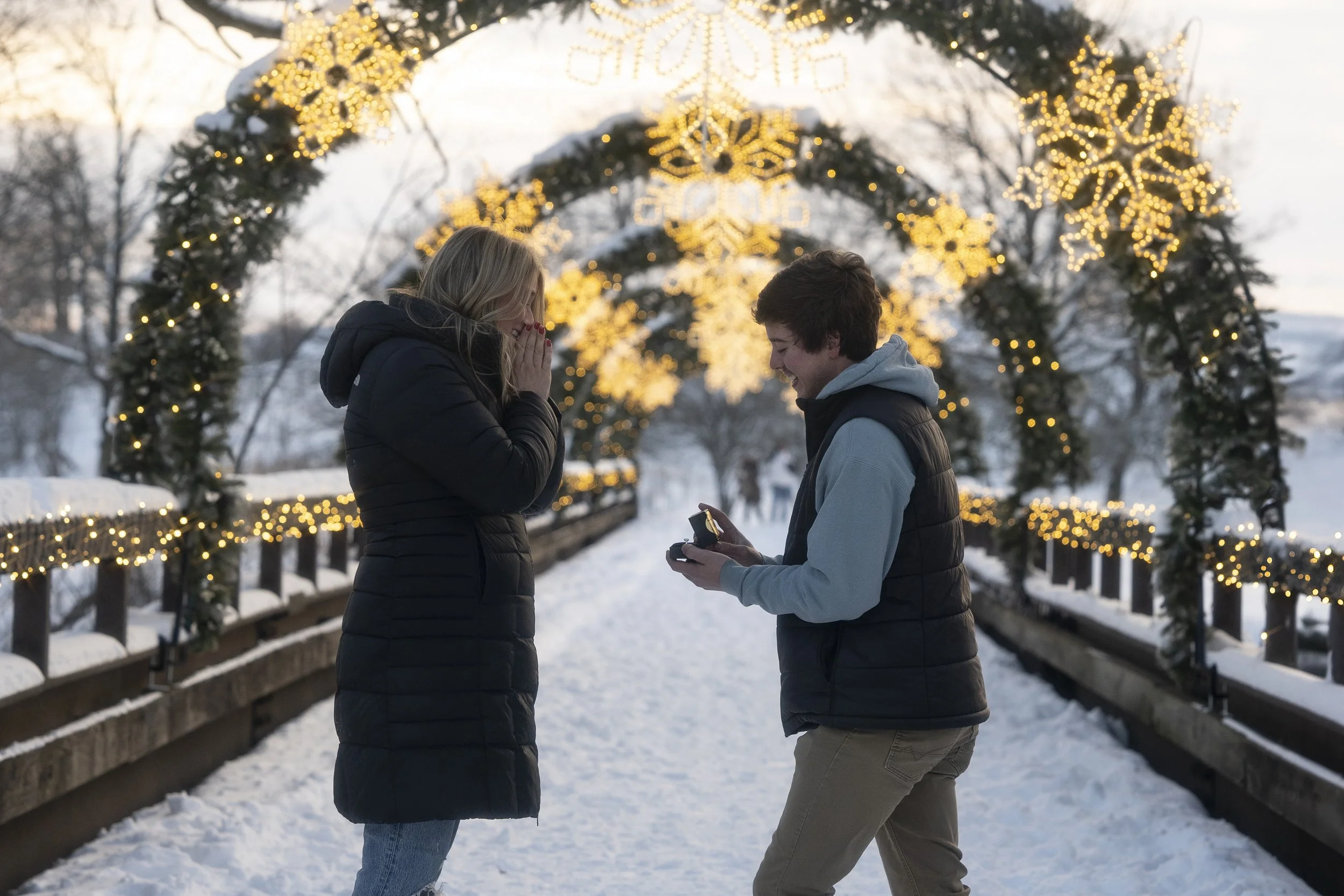 A man is proposing to a woman on a snowy bridge decorated with Christmas lights and greenery, with snow-covered trees in the background.