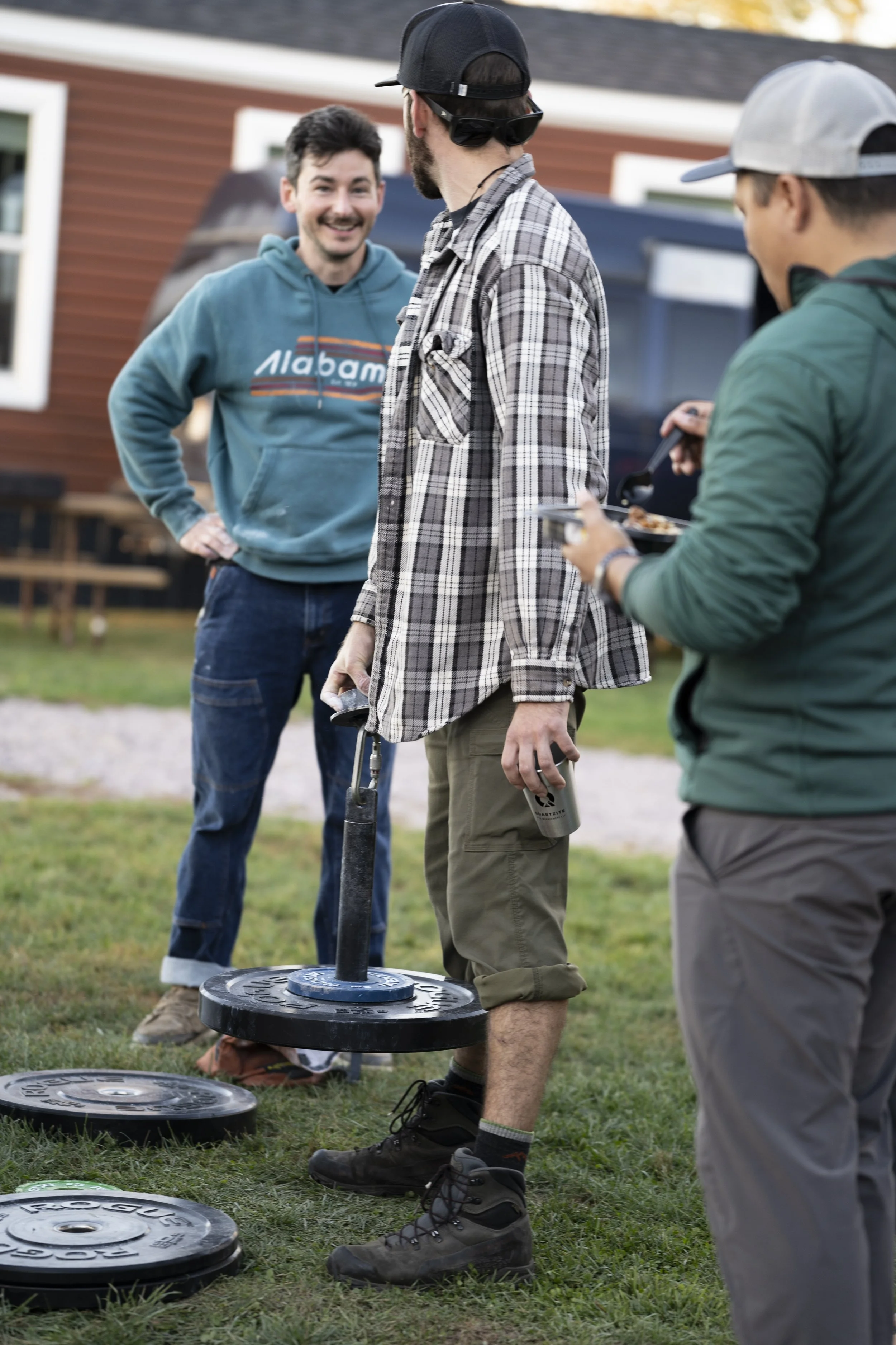 Three young men outdoors with a gym weightlifting plate on an outdoor grass area, one holding a weightlifting bar leaning on the ground, and two others talking and eating. One man is smiling, wearing a blue hoodie, another in sunglasses and a black c