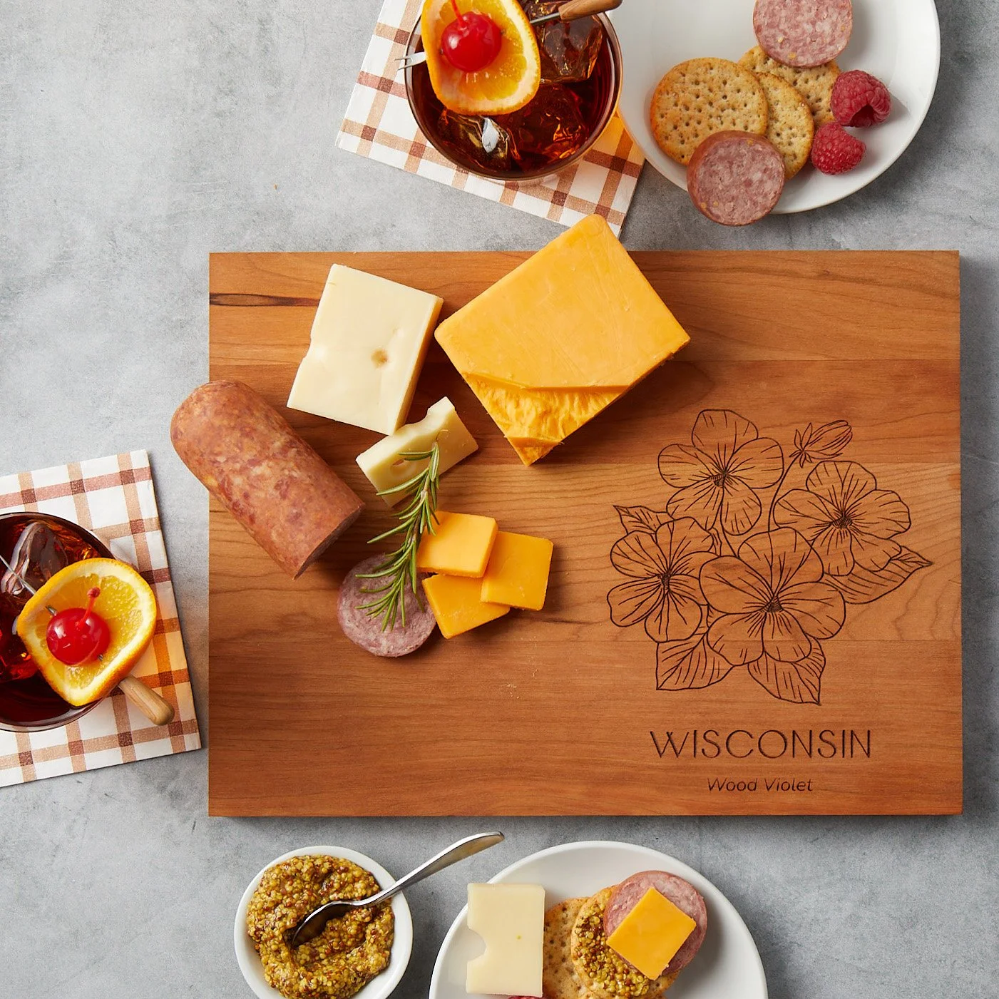 A Wisconsin state flower inscribed cutting board styled with cheeses, mustard, old fashions and sausage.
