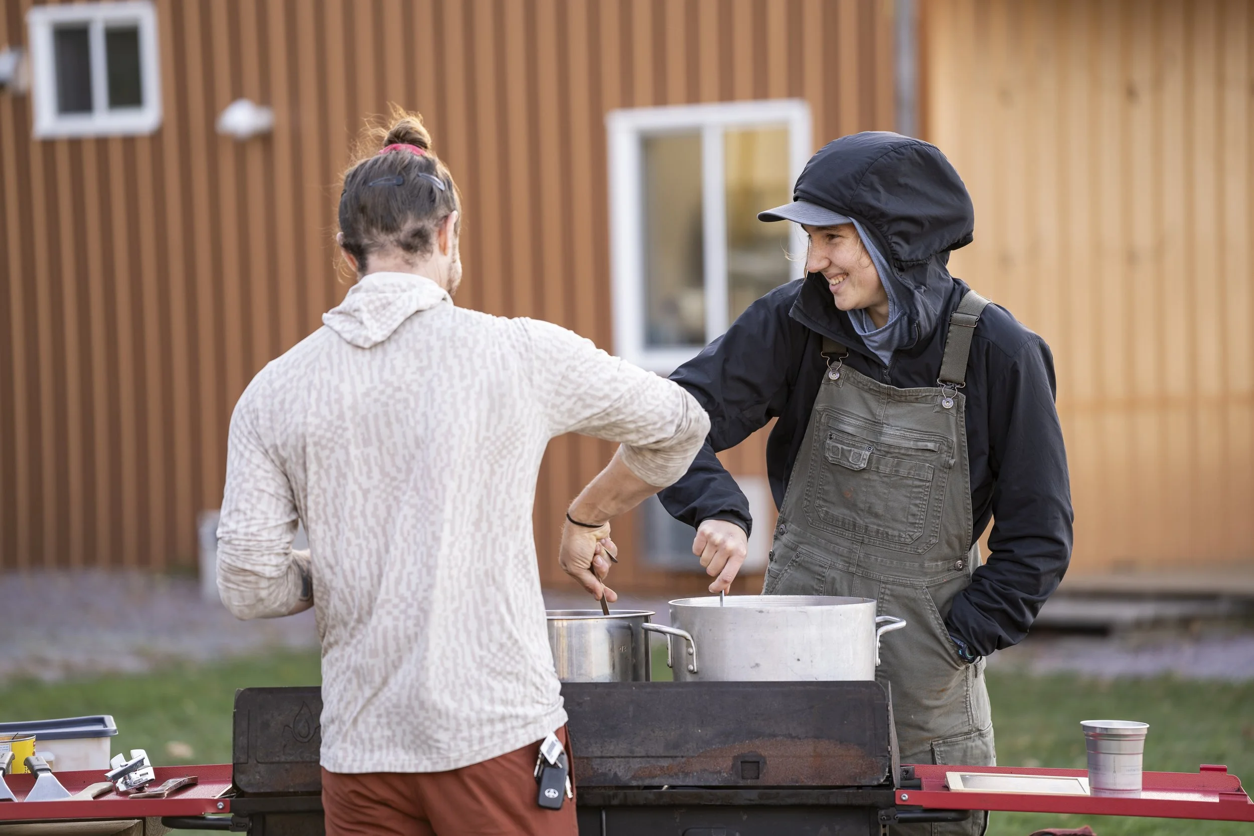 Two people are outdoors, laughing and serving food from pots at a barbecue grill, with a wooden building in the background.