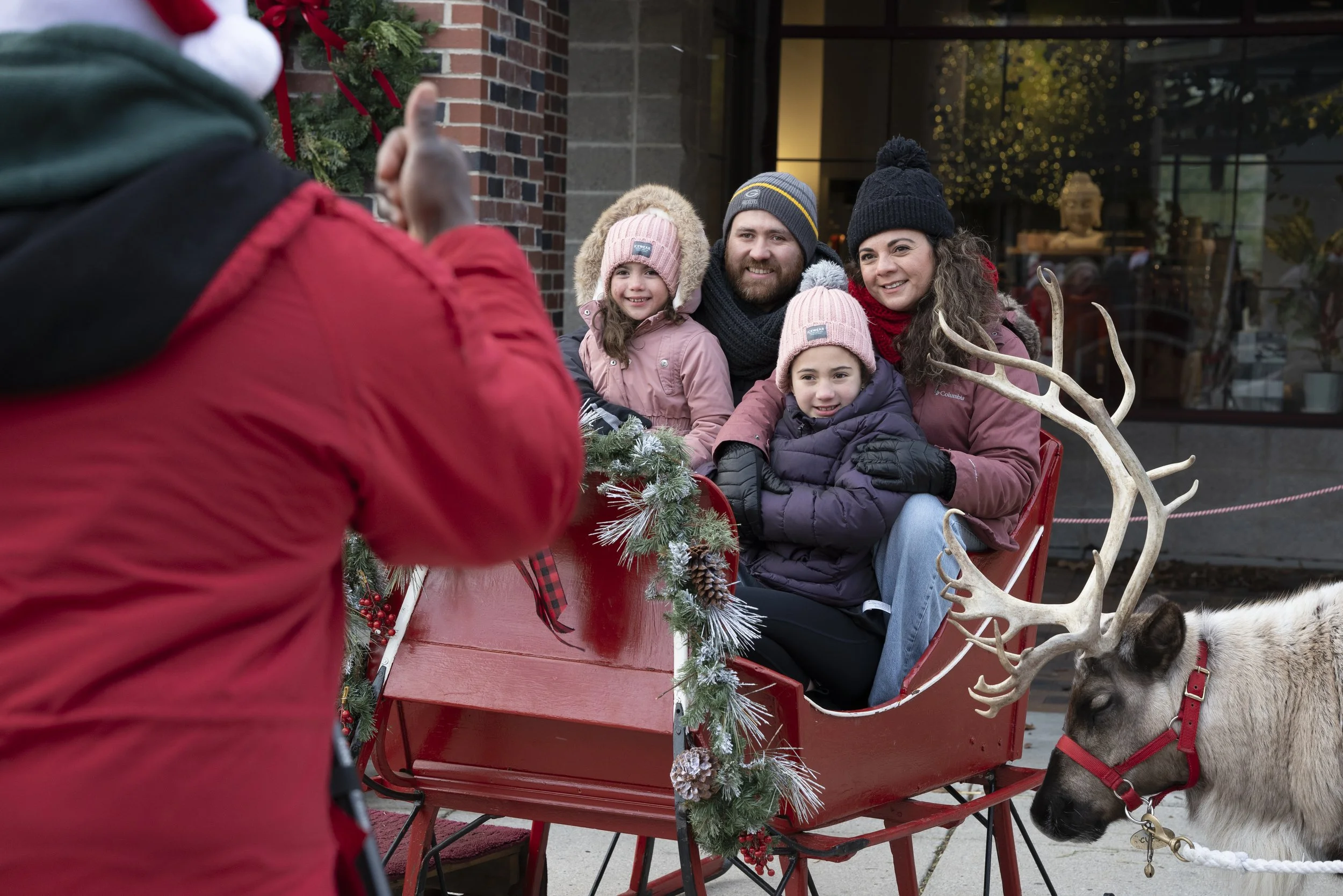 Family of four sitting in a red sleigh decorated with Christmas greenery, taking a photo with a person in a red jacket. A reindeer with antlers is nearby.