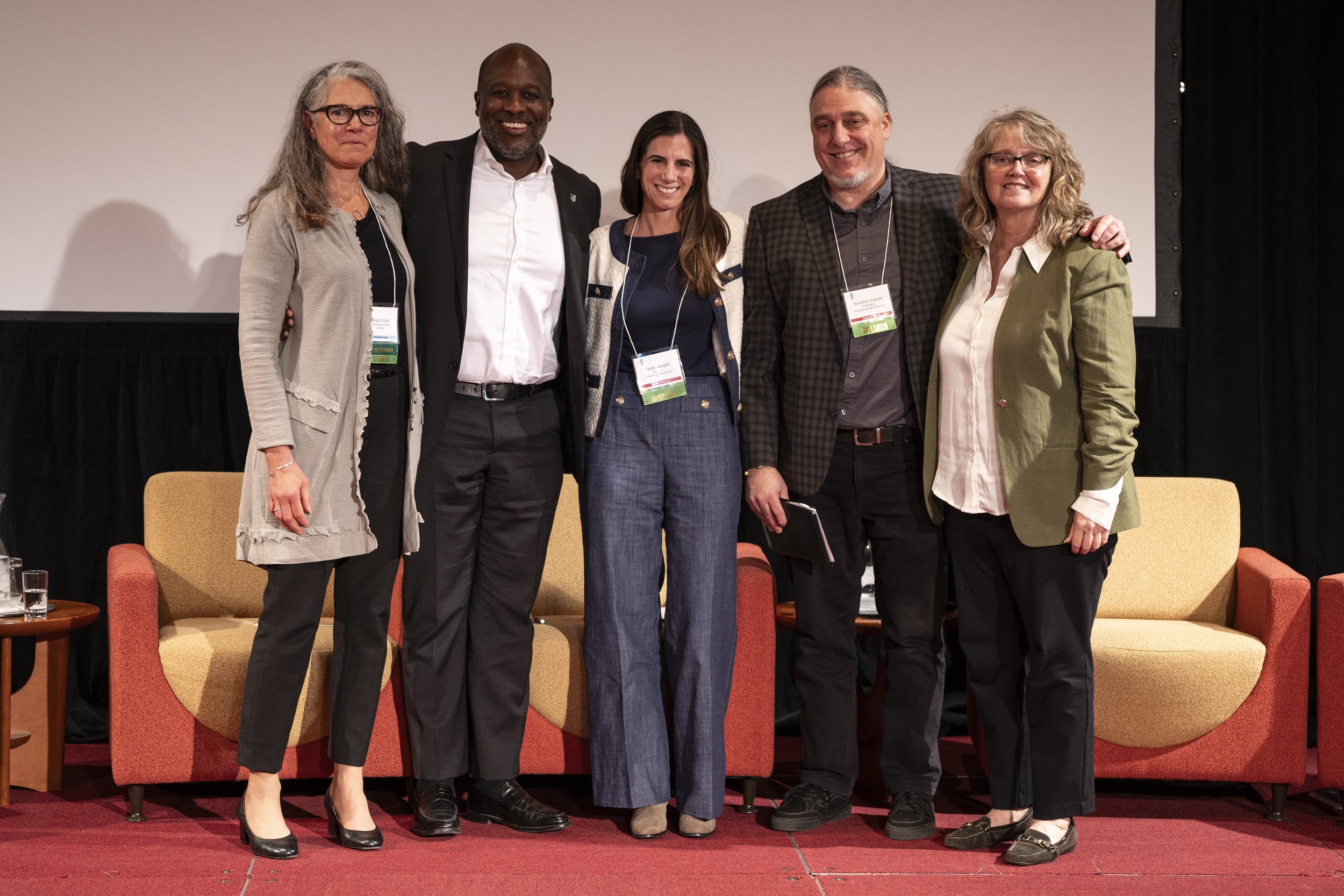 Six diverse people standing on a stage in front of a white screen, smiling, with three arm chairs behind them, at a conference or event.