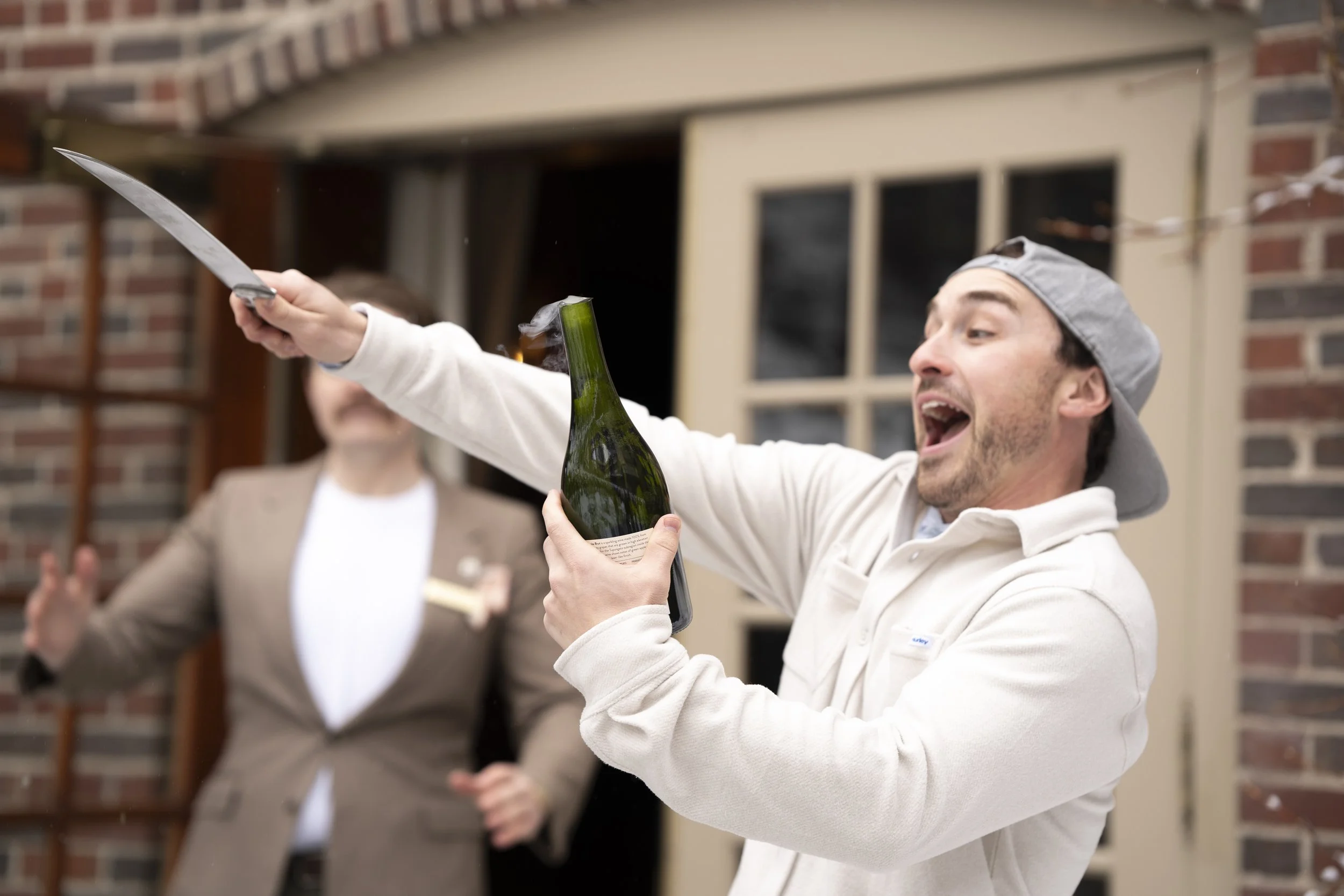 A man celebrating, holding a champagne bottle and a knife for cutting the bottle, with a man in a brown blazer smiling in the background. They are outdoors near a brick building.