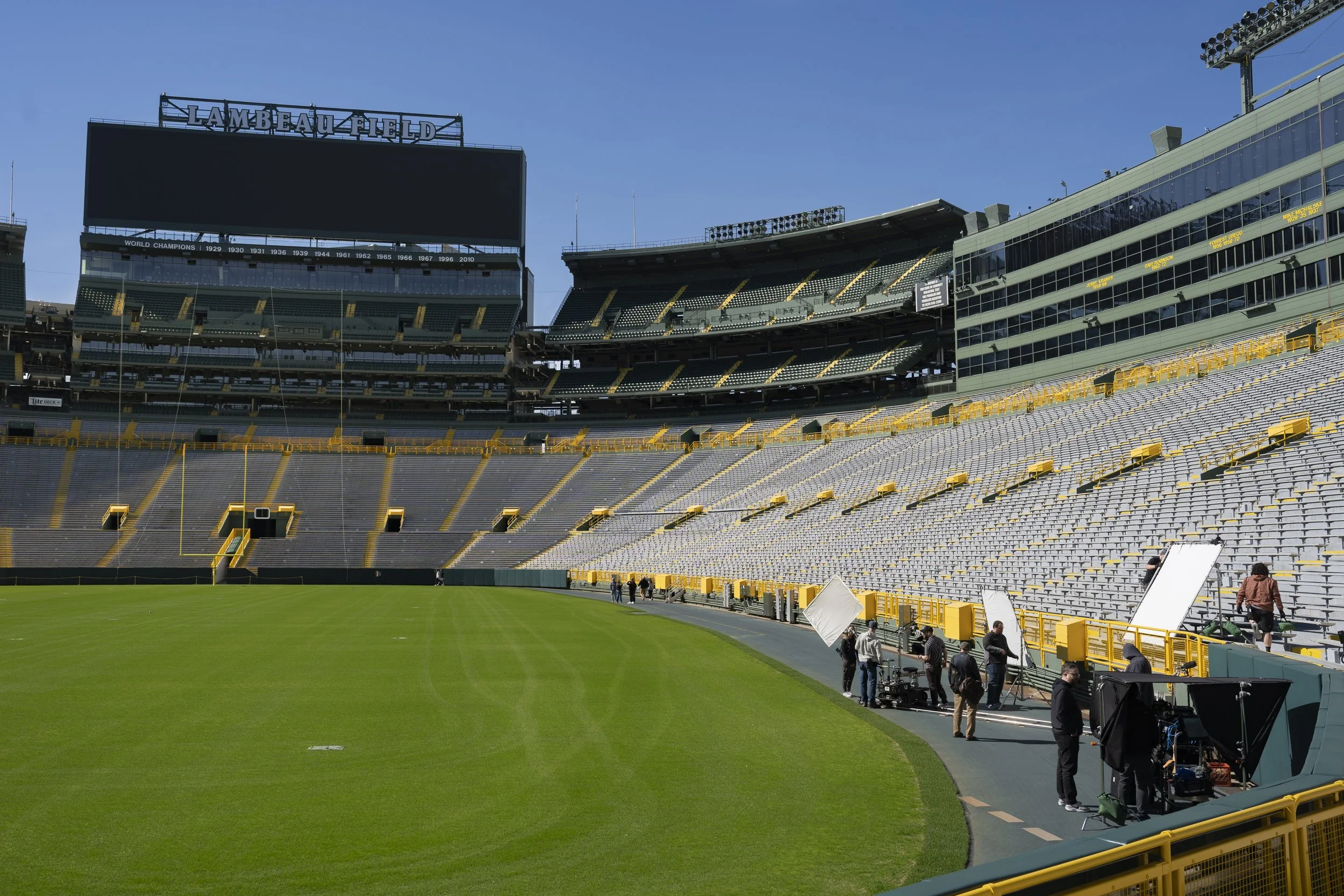 Empty sports stadium with crew members setting up equipment on the field, green grass, and yellow seating. Large digital scoreboard displaying "Lambeau Field," with a clear blue sky overhead.