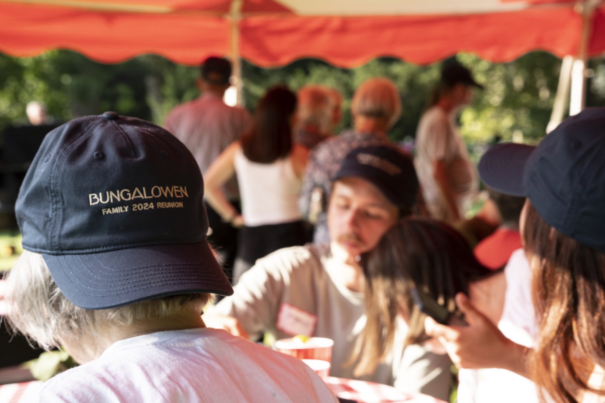 People gathered outdoors under a red canopy, some wearing dark baseball caps, at a family reunion event.