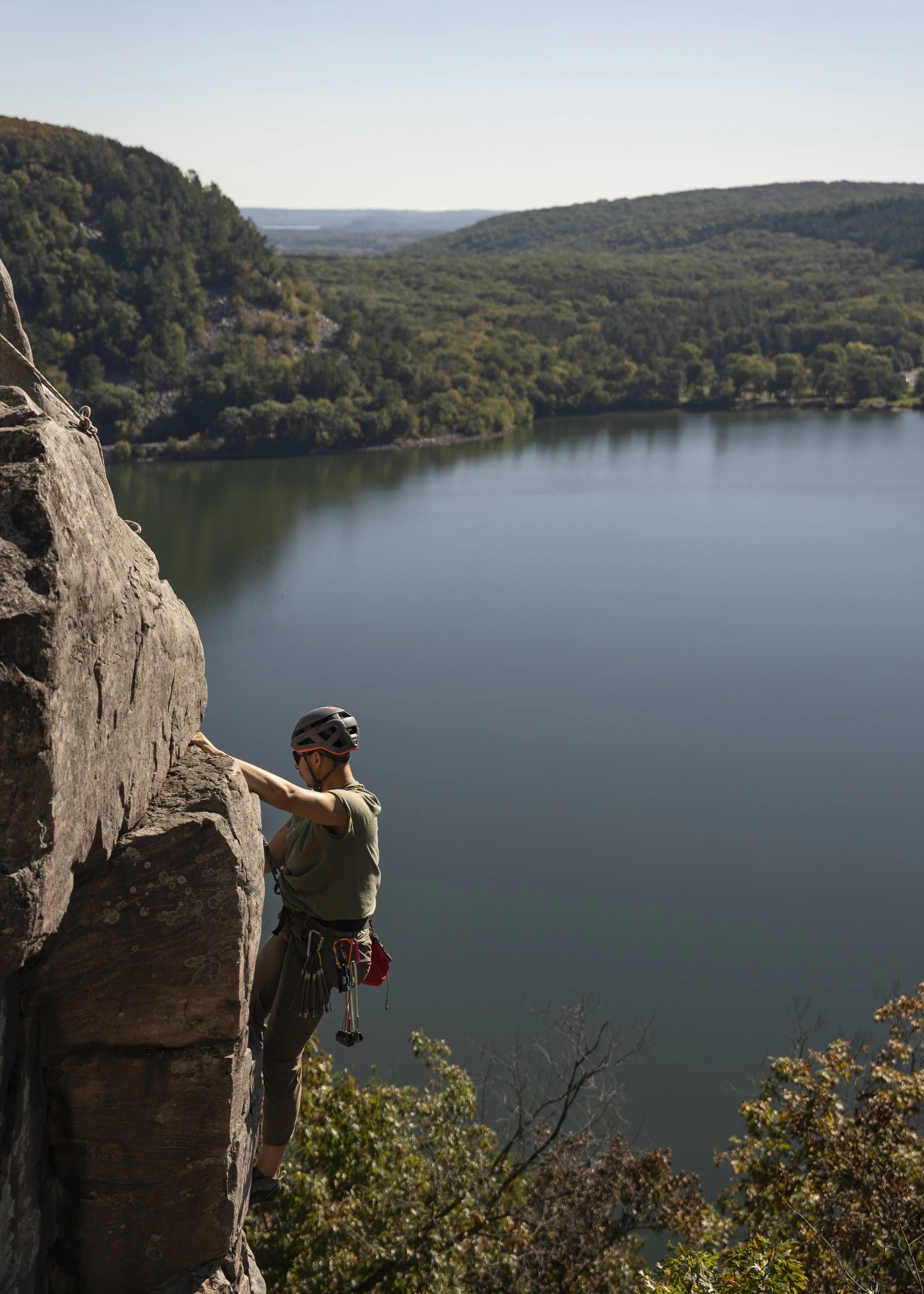 A person rock climbing on a cliffside overlooking a lake with green hills in the background.