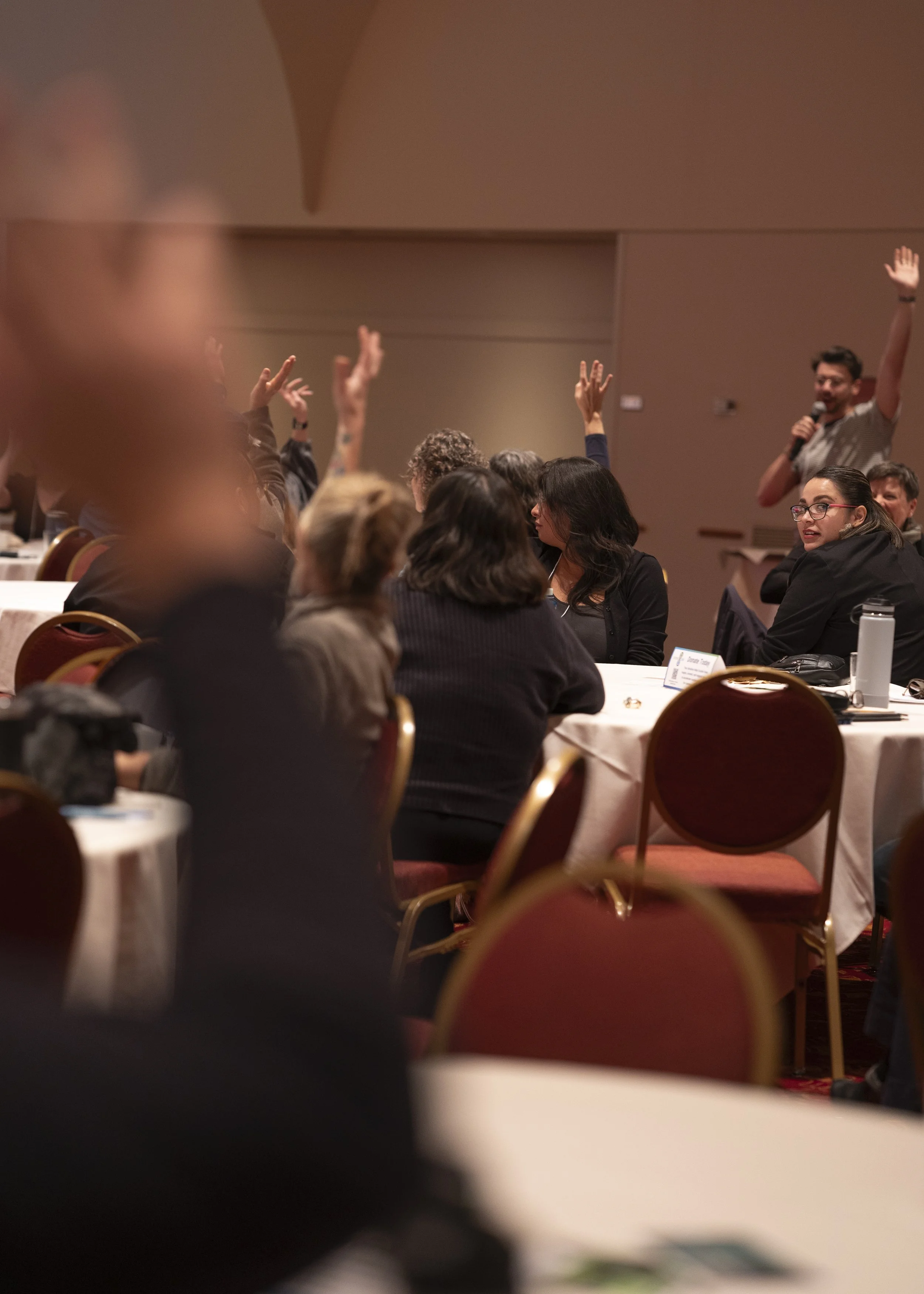 A group of people sitting at round tables in a conference room, with several raising their hands to ask questions or participate, and a person standing with a microphone in the background.