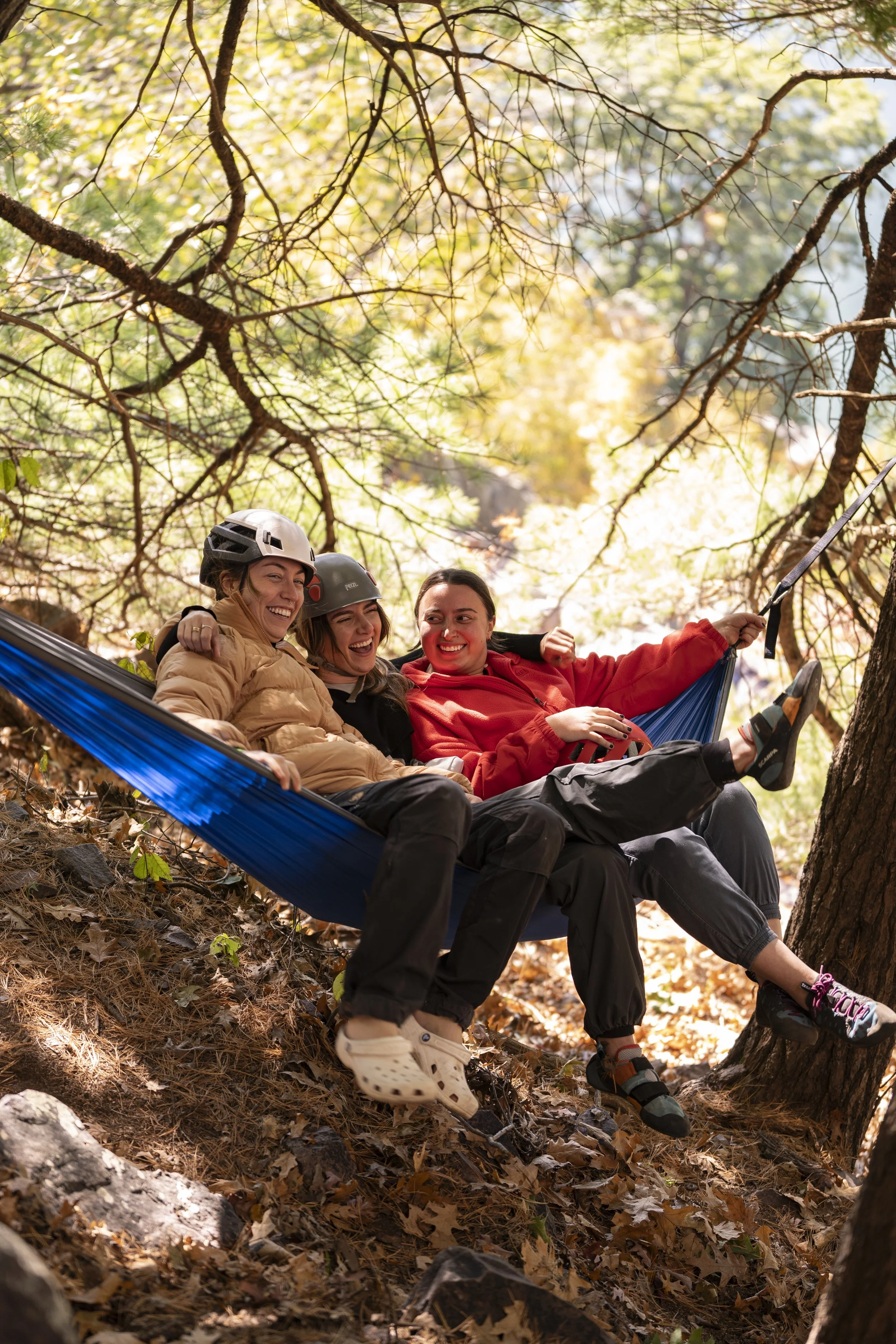 Three women sitting on a hammock hanging from trees in a forest, laughing and enjoying the outdoor scenery.