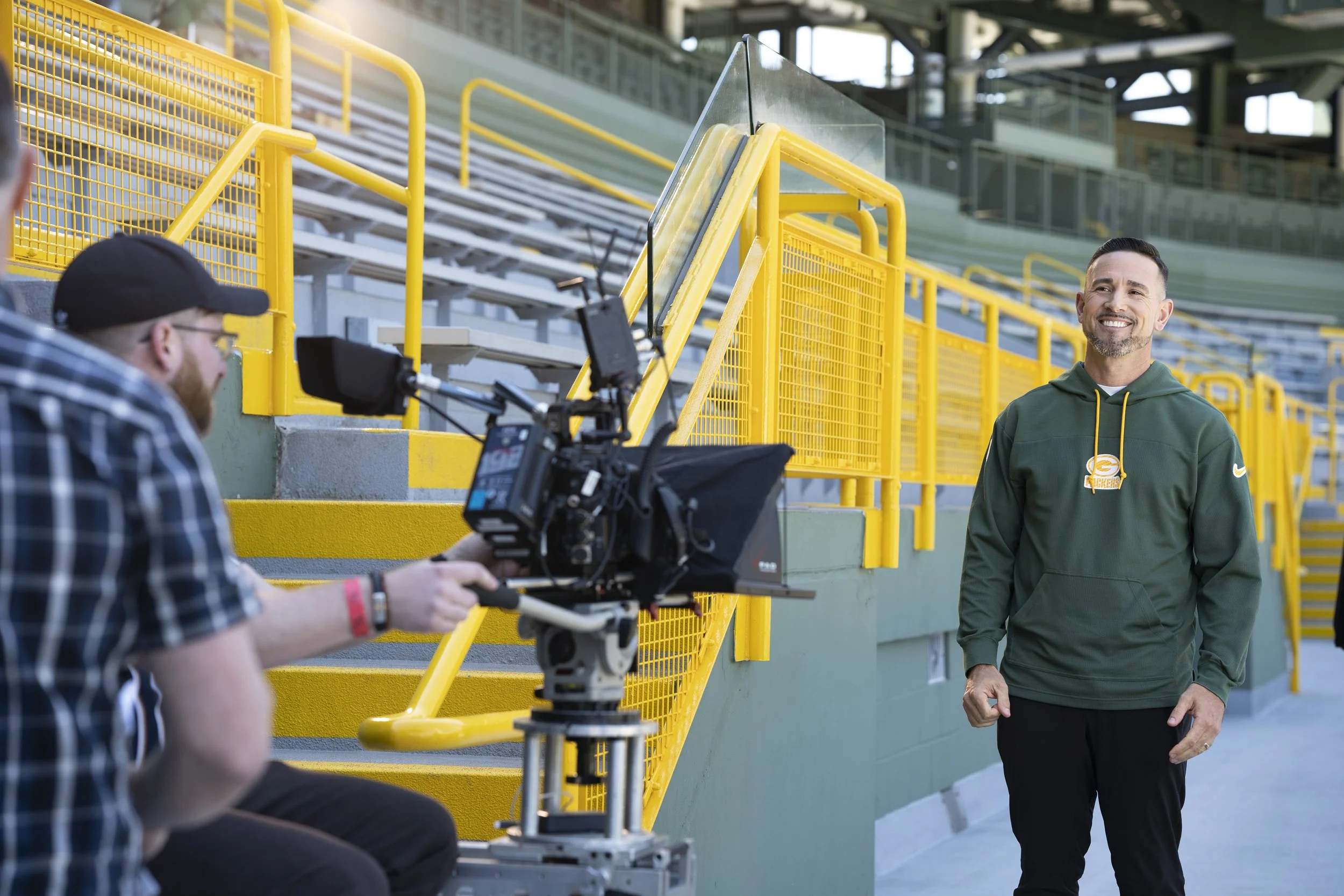 Man in green hoodie filming a man with a camera at a stadium with yellow railings.