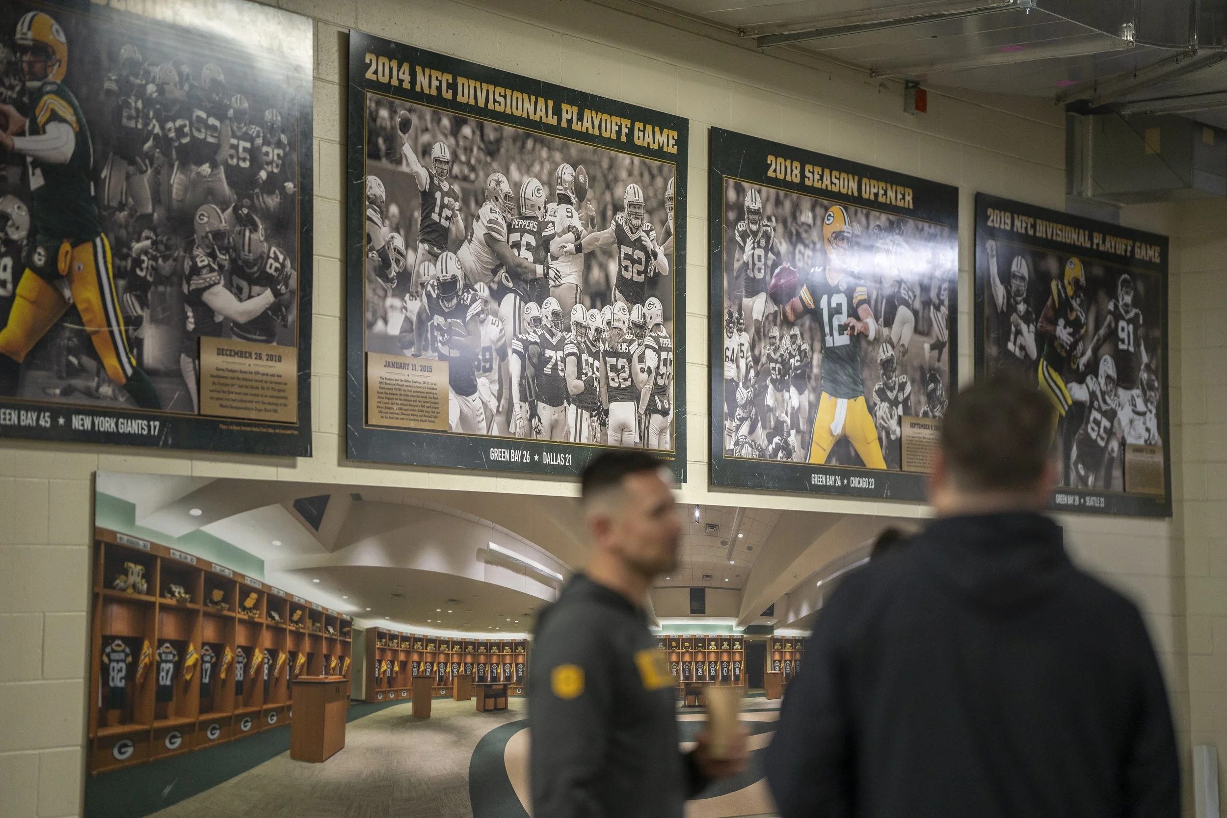 Two men talking in front of a wall with framed football photos and history, and a locker room with jerseys and helmets in the background.