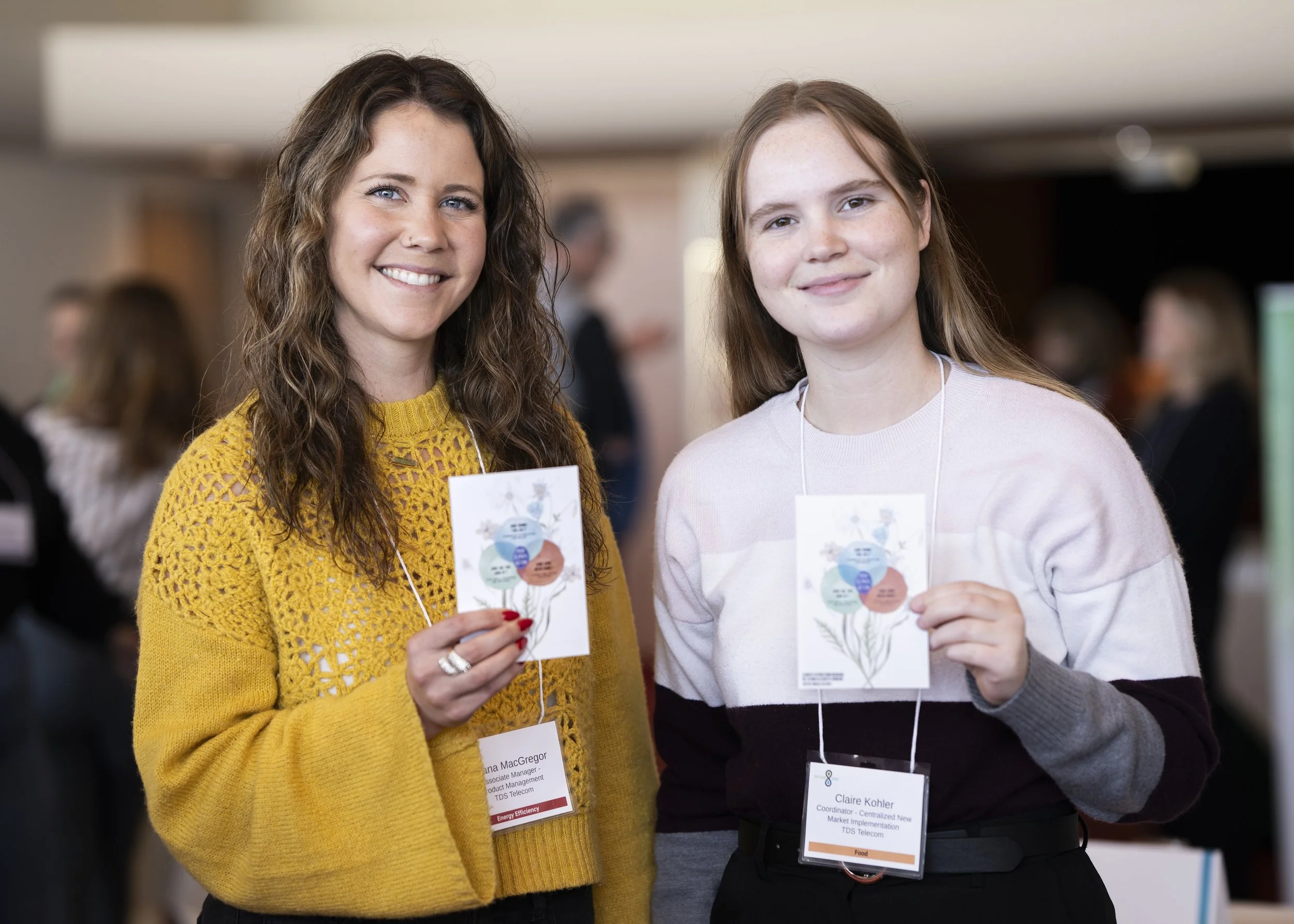 Two women standing side by side at a conference, holding a brochure and wearing name tags.
