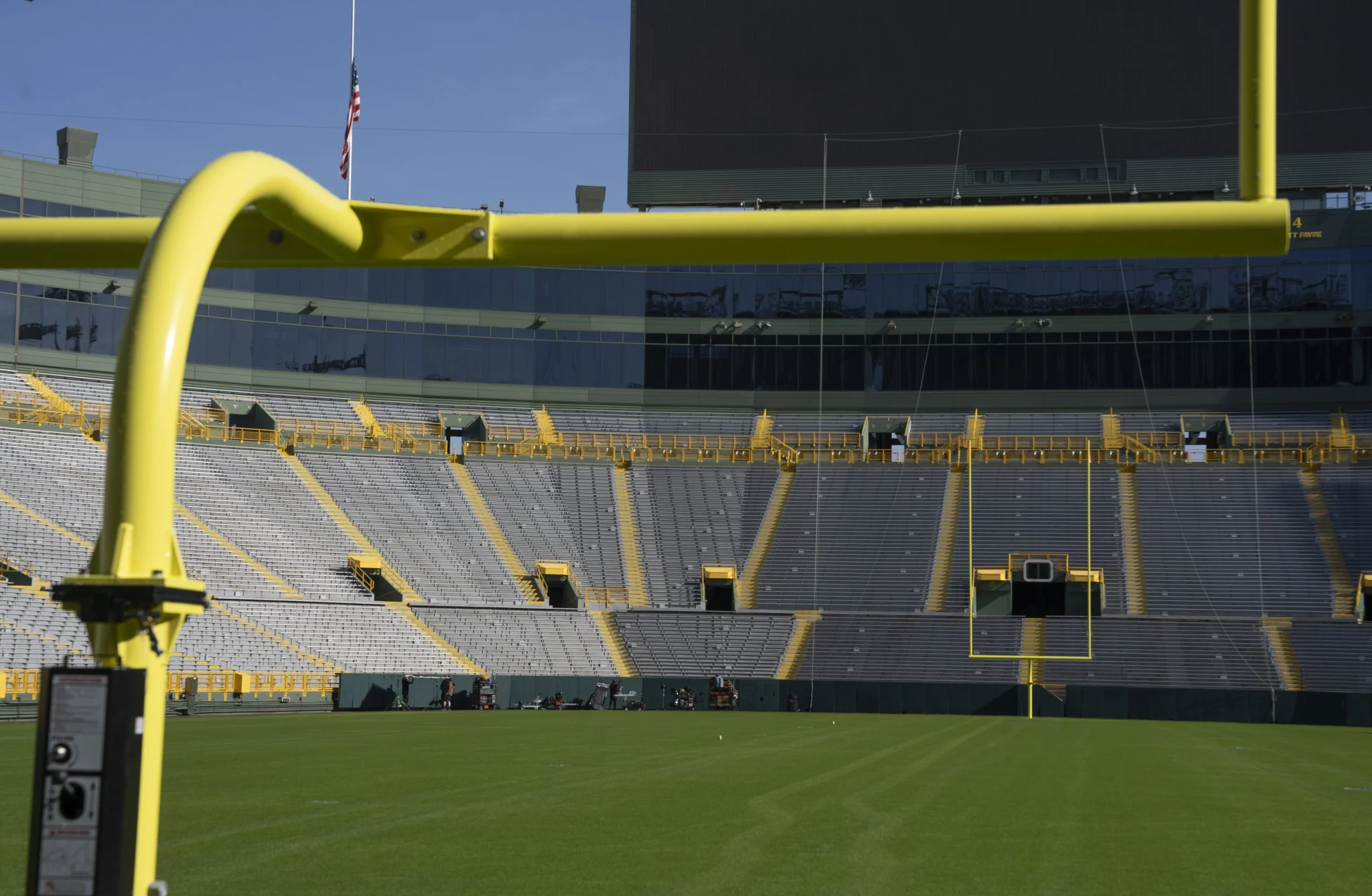Football field with goalpost in the foreground, empty stadium seating, and a large video screen in the background under a clear blue sky.