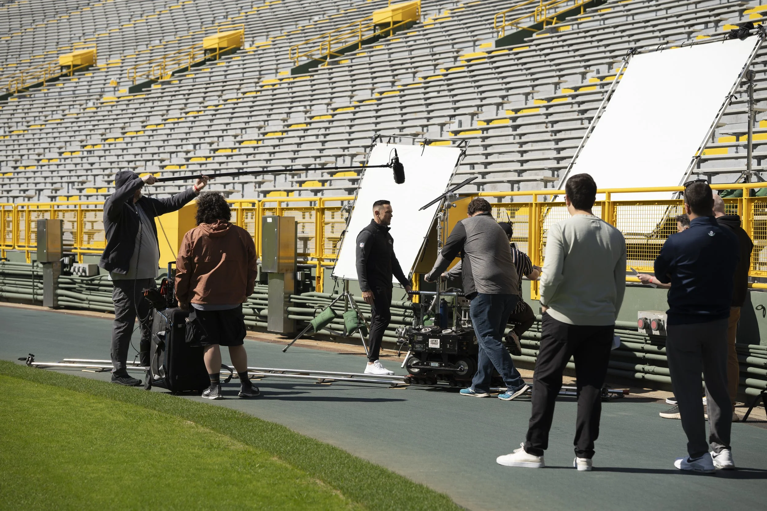 Film crew filming a scene on a sports field with yellow stadium seating in the background. Crew members are setting up equipment and a large white reflector or screen.