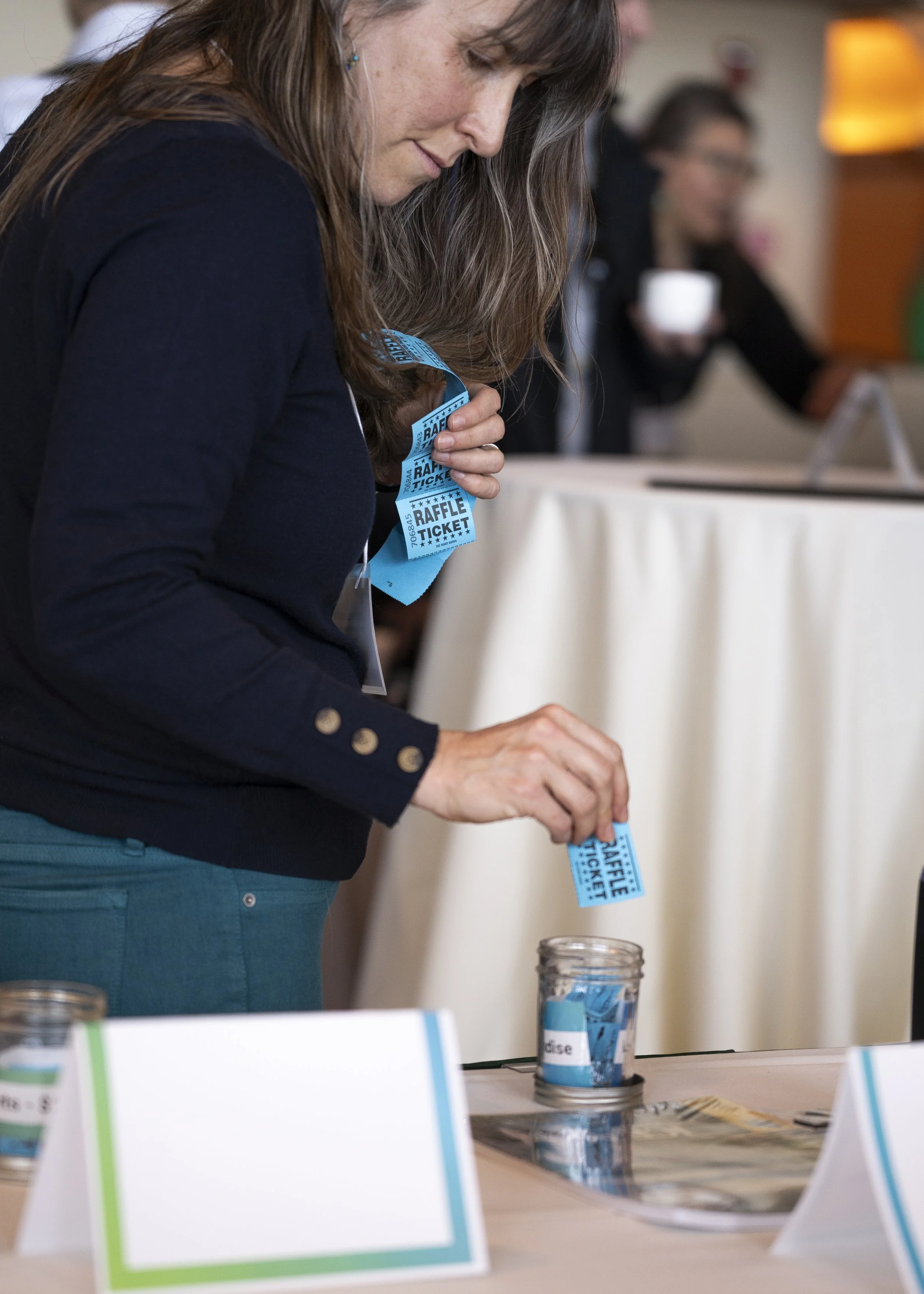 A woman with long brown hair wearing a navy sweater is placing raffle tickets into a glass jar at a registration or event table.
