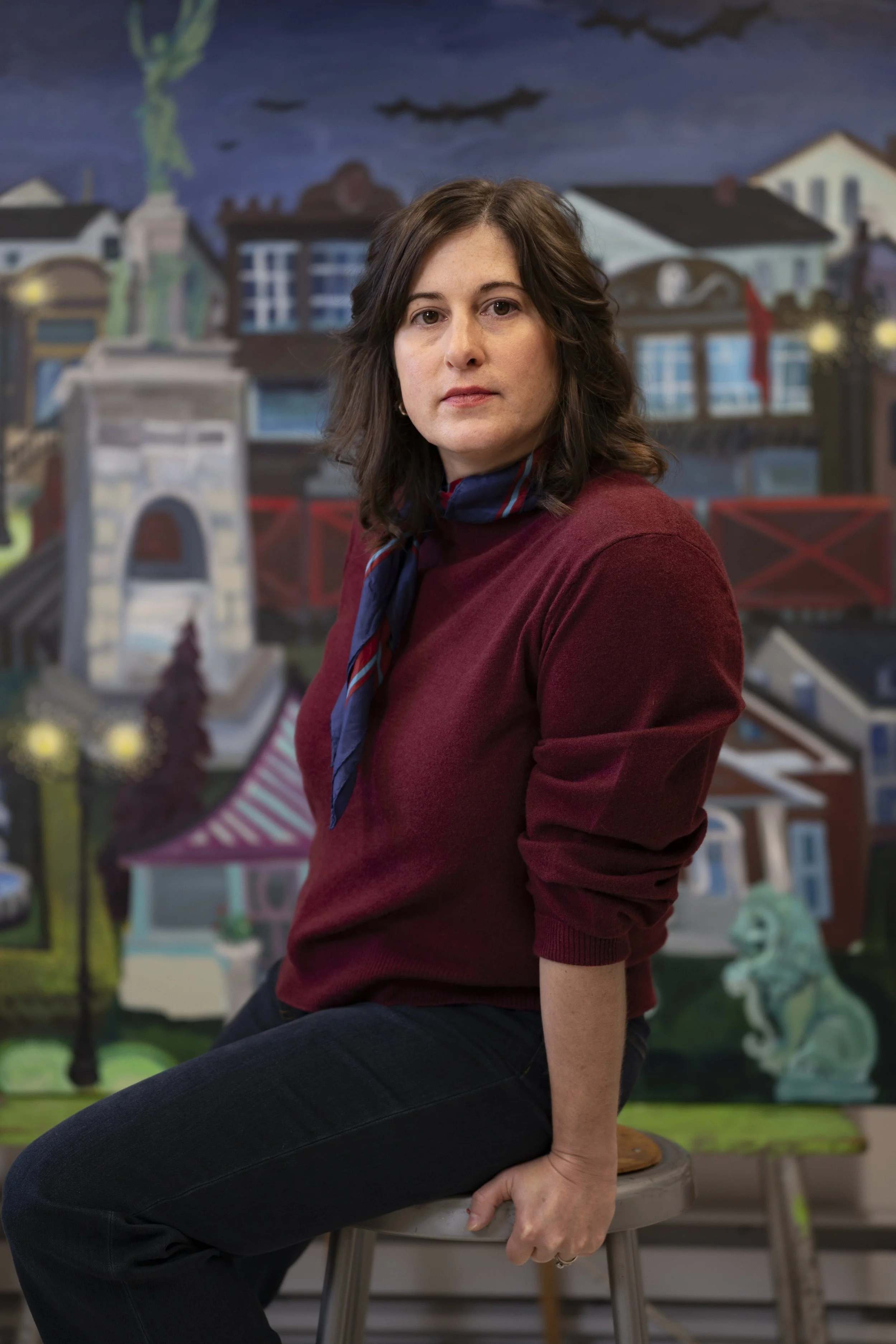 A woman with shoulder-length brown hair wearing a maroon sweater and a blue and red scarf, sitting on a stool in front of a colorful mural depicting a town scene with bridges, buildings, and a lion statue.