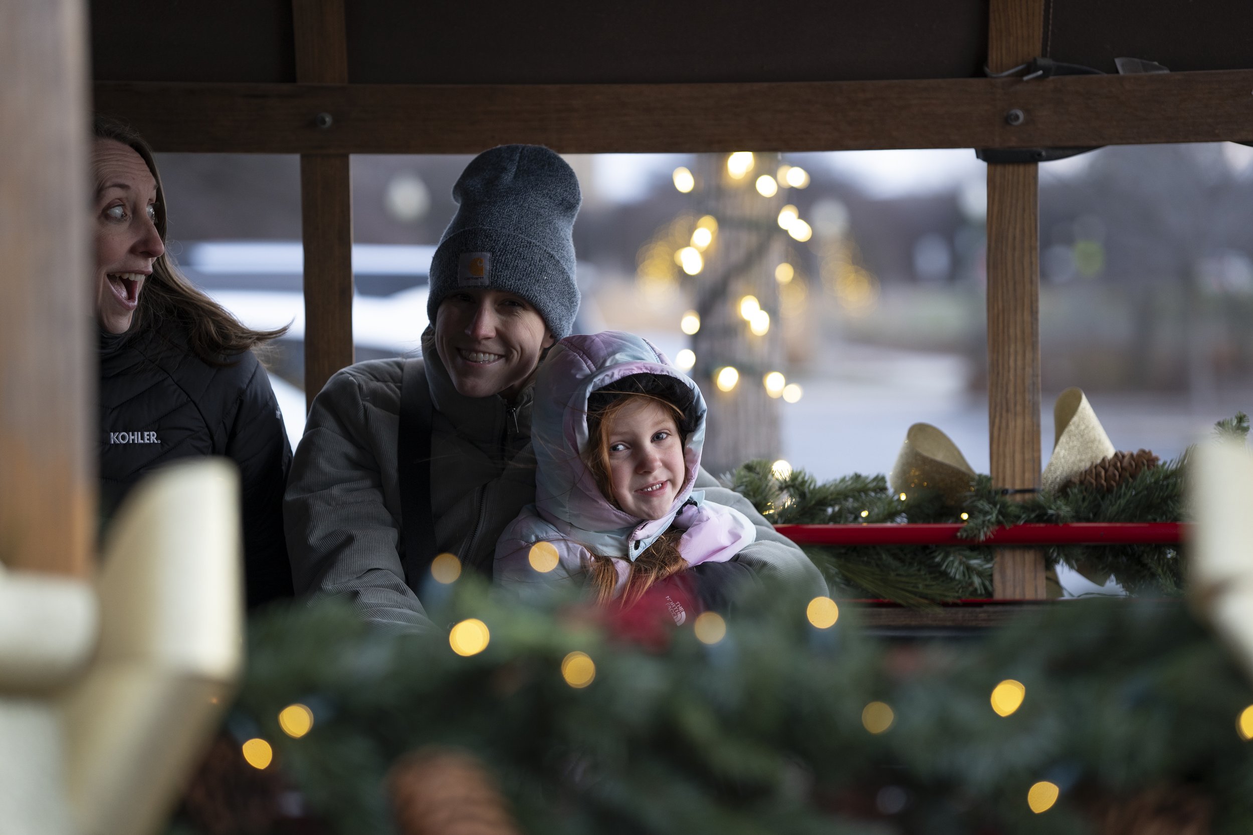 A family of three, a woman, a man, and a young girl, smiling and enjoying holiday festivities under a wooden structure decorated with Christmas greenery and lights.