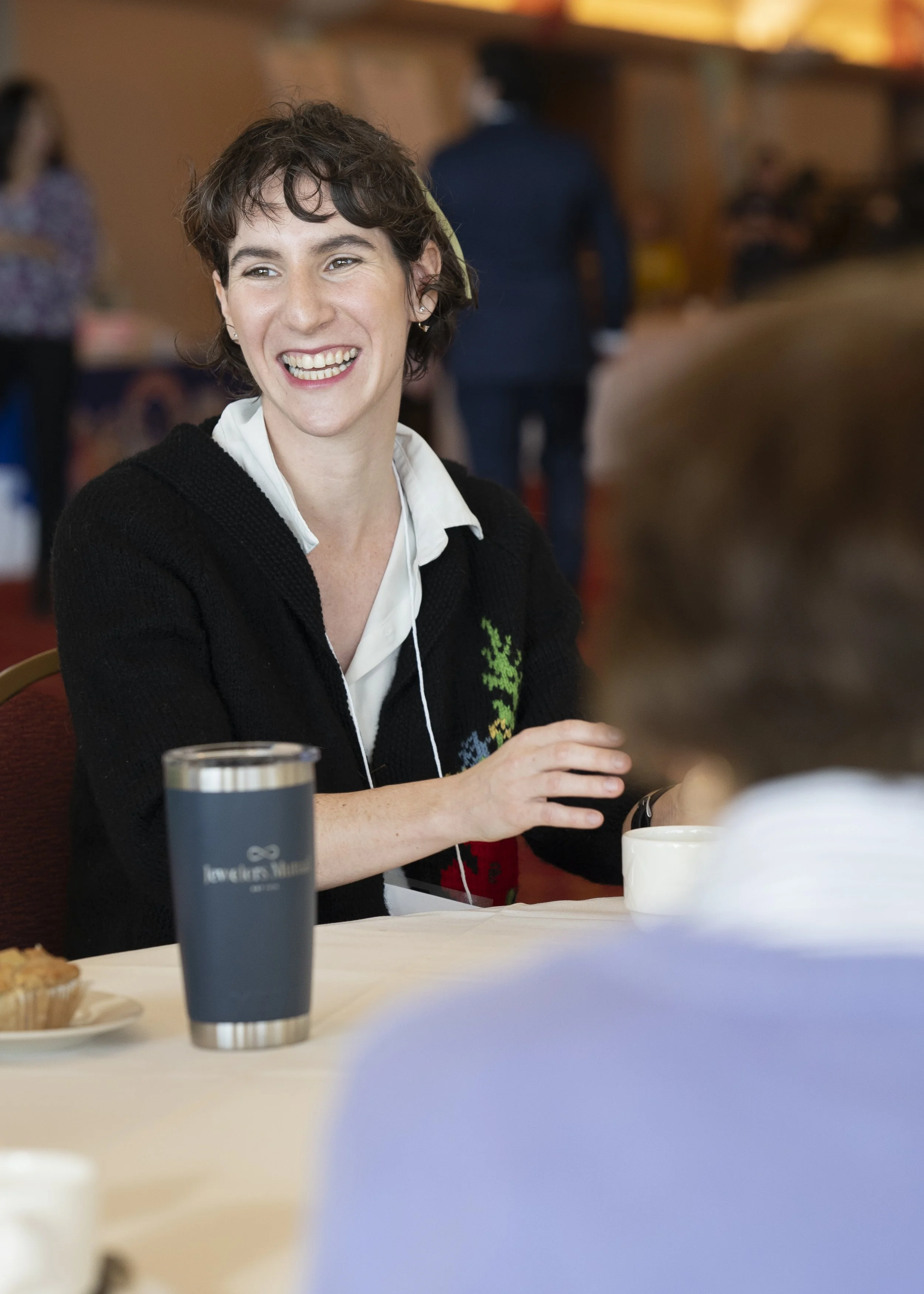A woman with short curly brown hair, wearing a white collared shirt and black sweater with colorful embroidery, smiling and sitting at a table during a social meeting or conference.