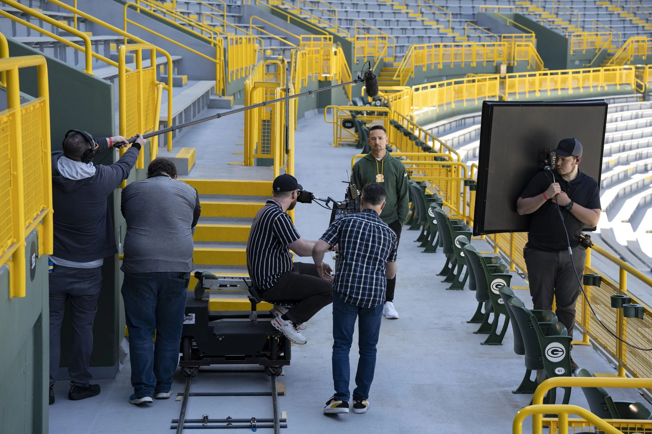 Filming crew setting up a scene in an empty sports stadium with green seats and yellow railings. Crew members are placing equipment, adjusting lights, and preparing a shot with Matt LaFleur in a green hoodie.