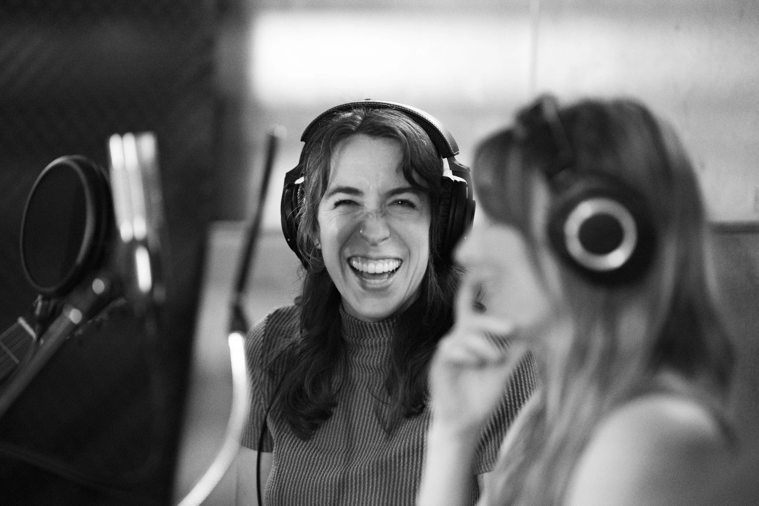 Two women in a recording studio, wearing headphones, laughing and speaking into a microphone.