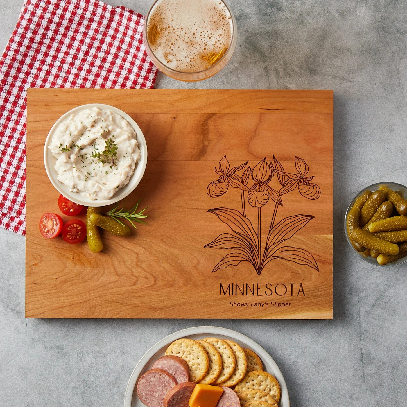A Minnesota state flower inscribed cutting board styled with some spread, a beer, cheese, sausage and crackers and a jar of pickles. 