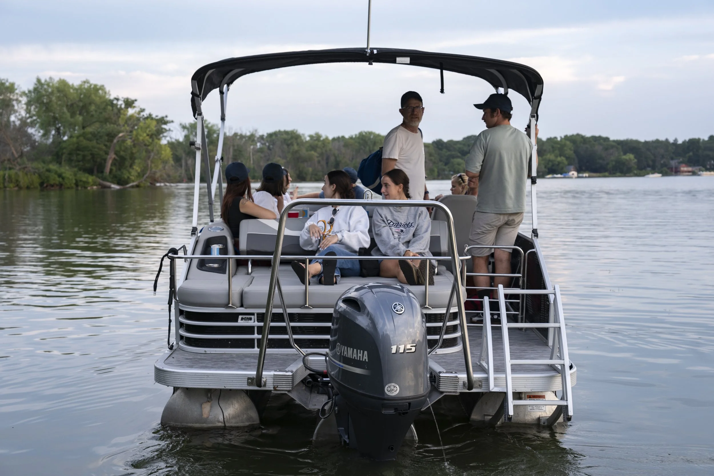 People on a pontoon boat on a calm river with trees in the background during dusk.