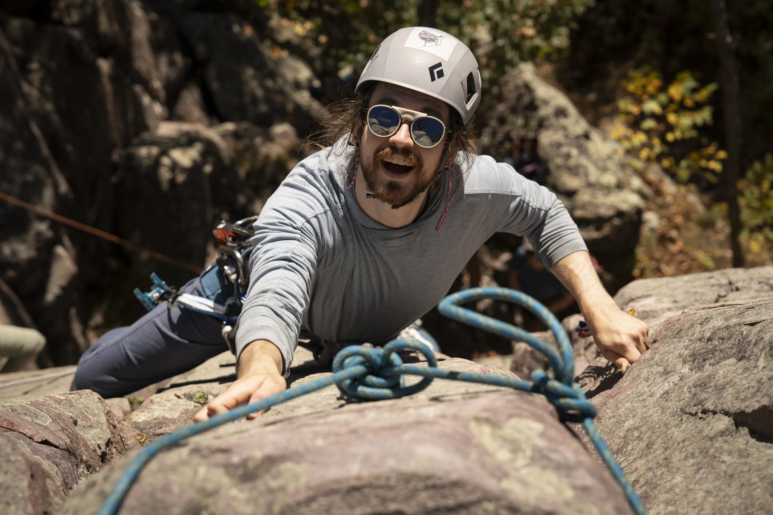 A man climbing a rock face outdoors with a climbing rope, wearing a helmet and sunglasses, smiling.