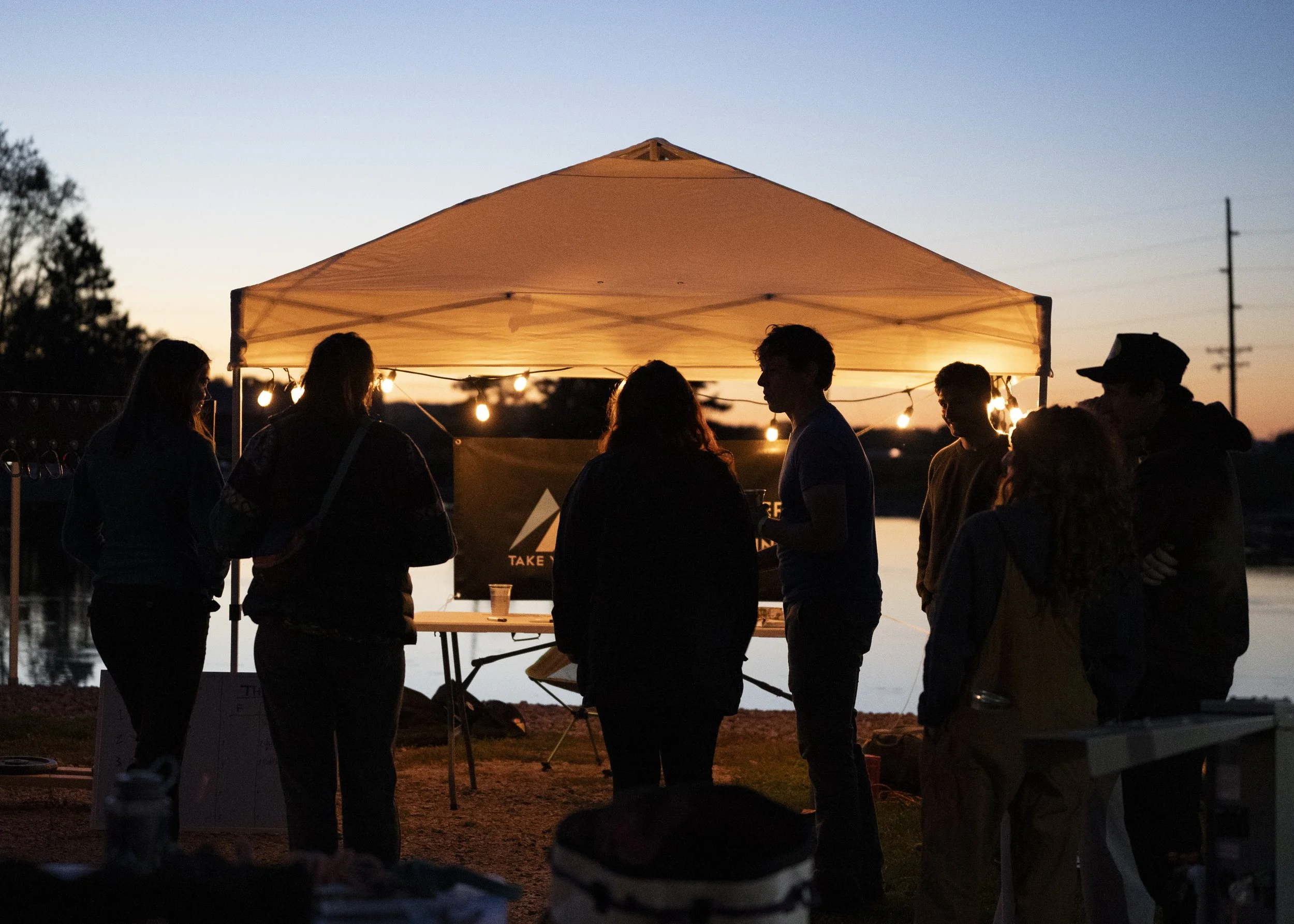 Group of people gathered under a lit canopy near a body of water during sunset.