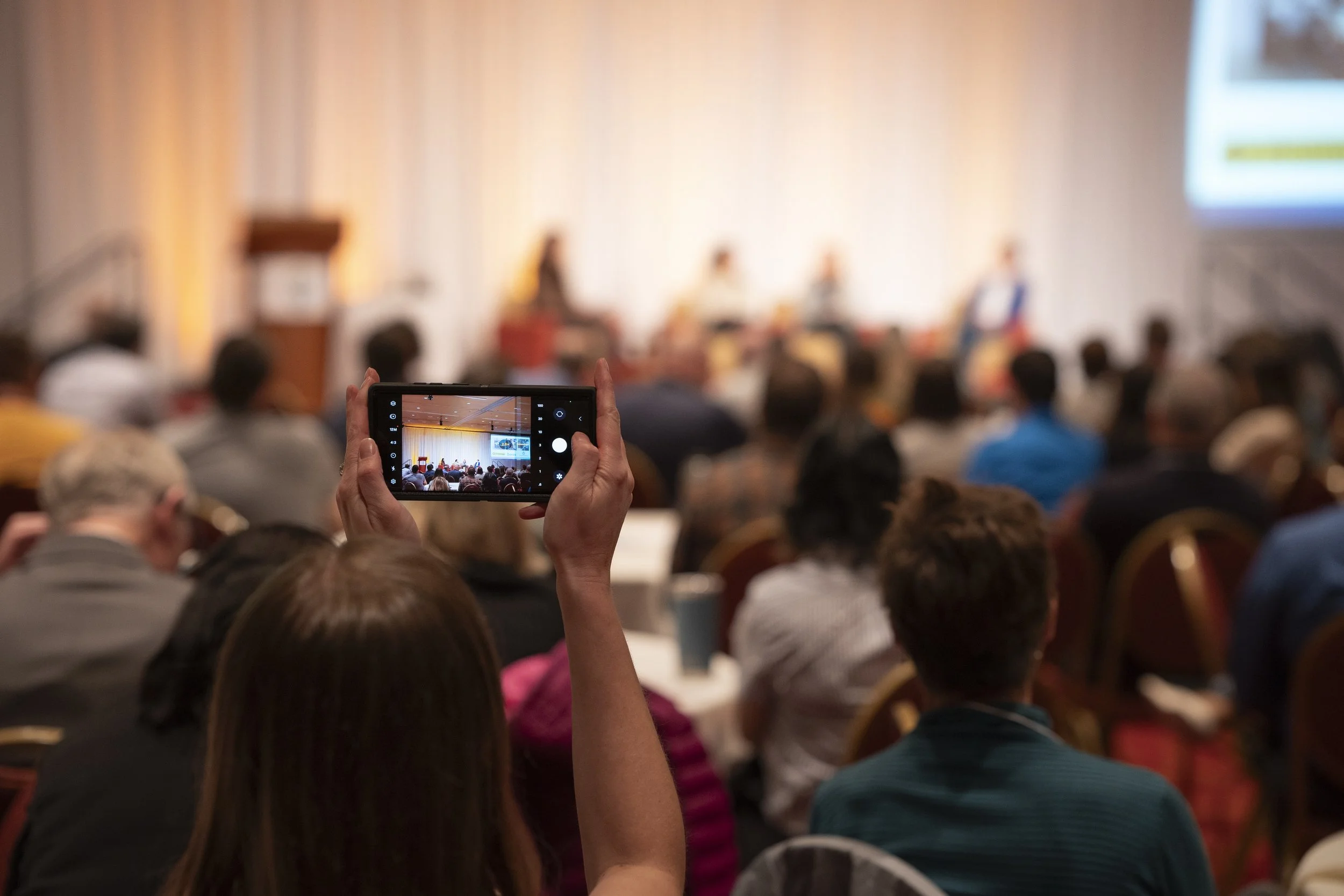 Person taking a photo or recording a video of a conference or panel discussion on their smartphone, with a blurred audience and stage in the background.