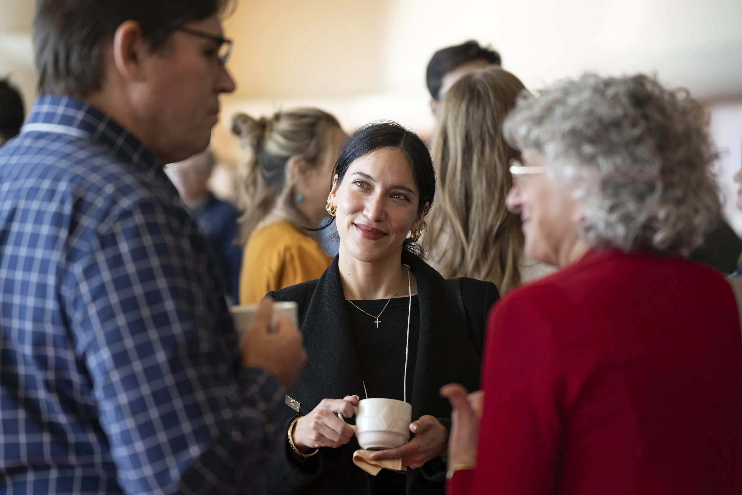 Group of people at a social event, engaging in conversation, woman in black holding a coffee cup, smiling, wearing gold jewelry, woman in red with gray hair and glasses, man in blue plaid shirt.