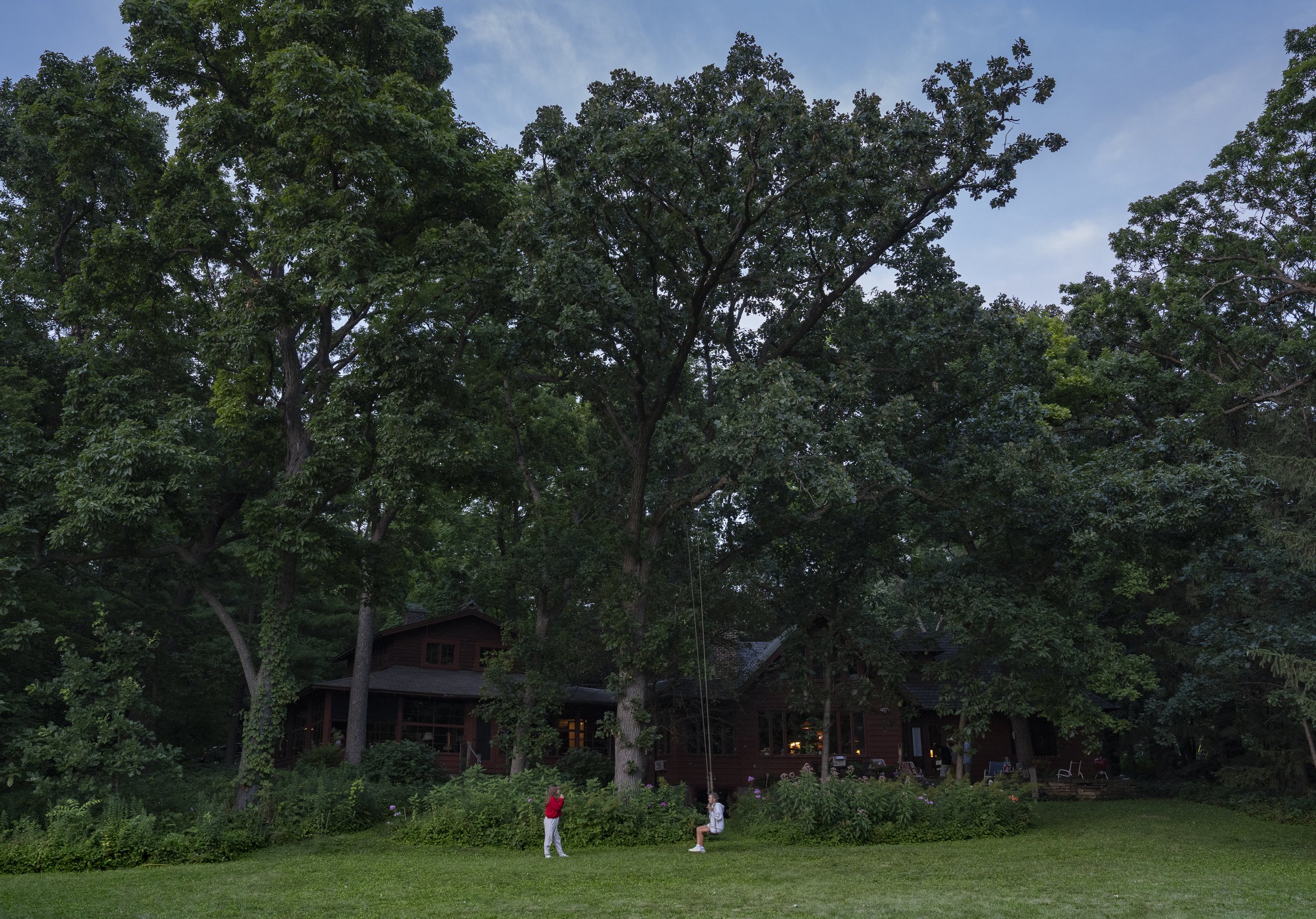 A large house surrounded by tall trees, with two children playing on swings in the front yard and a well-manicured lawn.