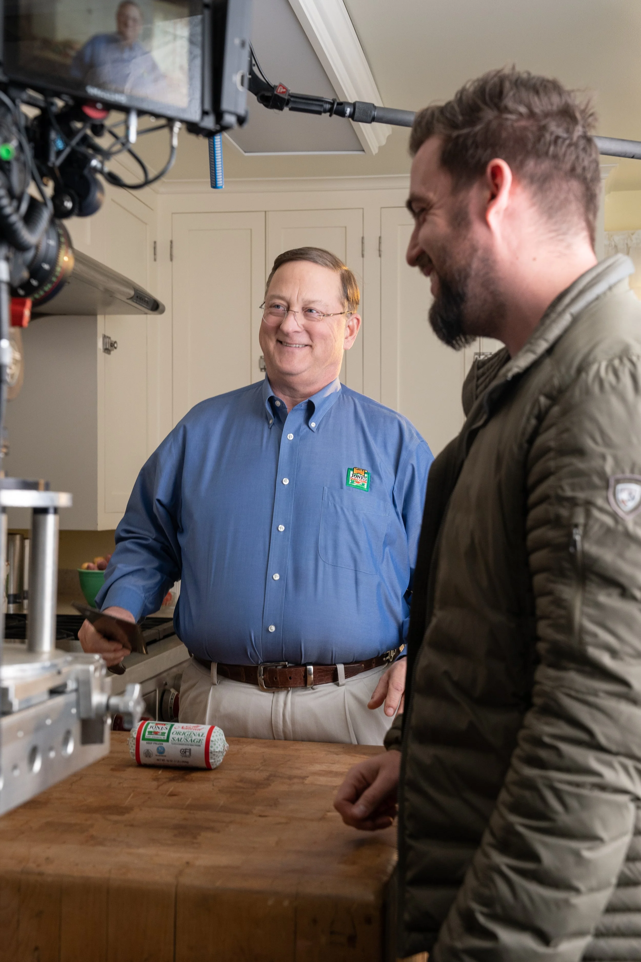 A man in a blue shirt and glasses smiling at another man in a brown jacket while filming on a camera, in a kitchen setting with a wooden countertop and a package of sausage.