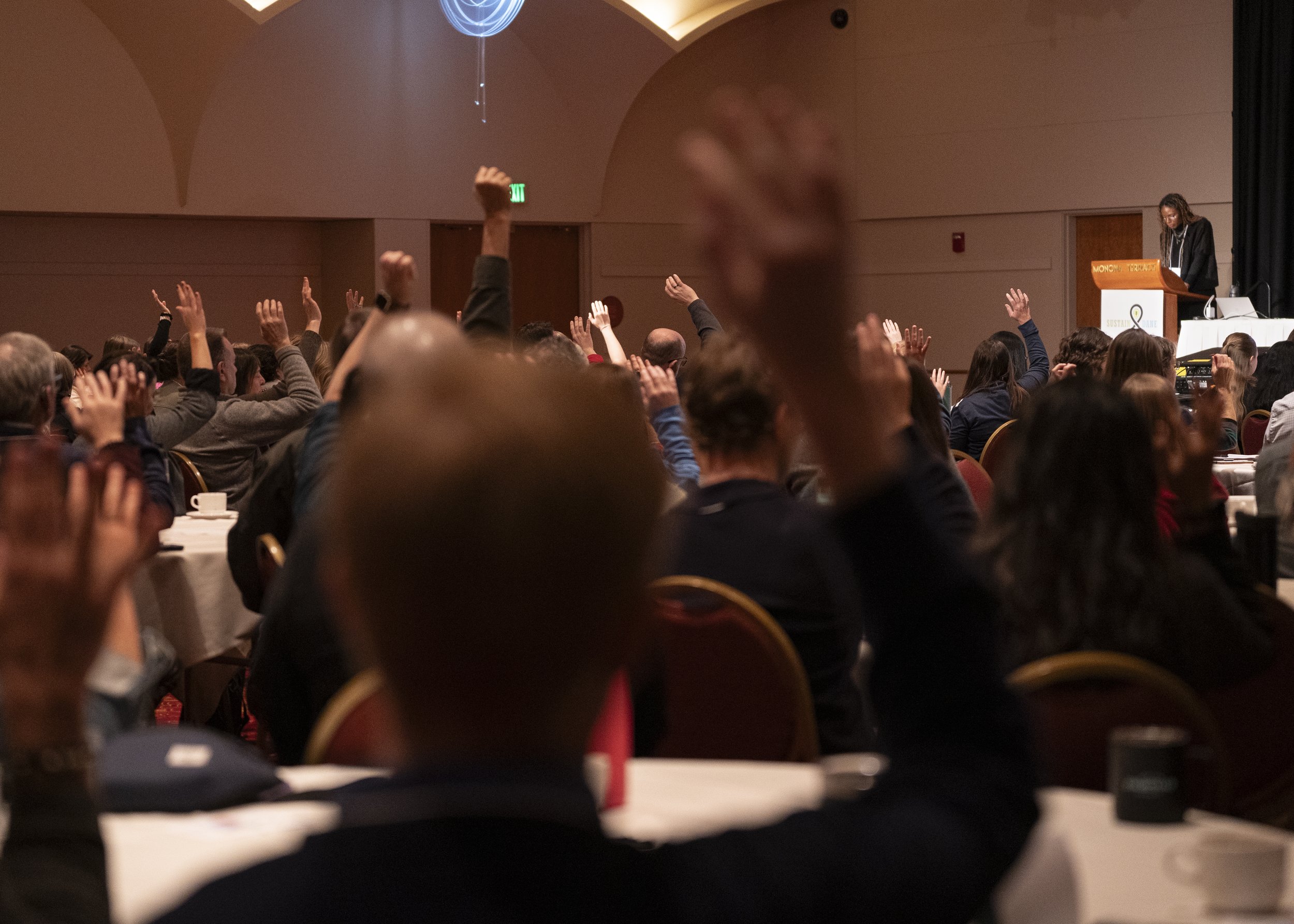 Audience members at a conference with many raising their hands while a speaker stands at a podium on stage.