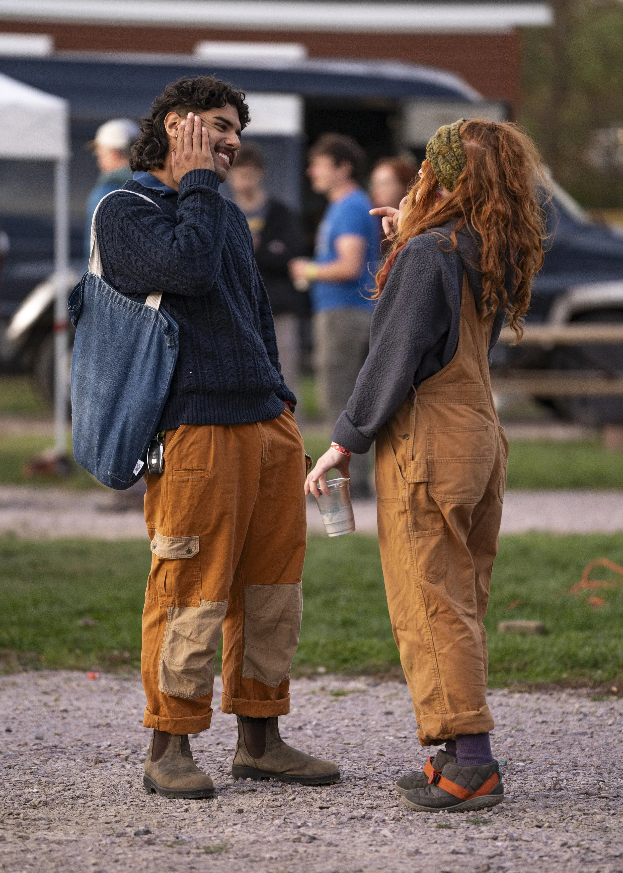 Two people standing outdoors, smiling and talking; one has a denim bag, the other holds a clear cup with a drink; group of people and tents in the background.