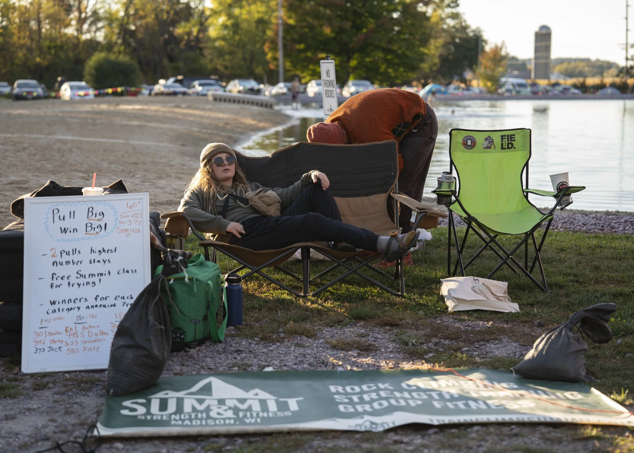 A person lounging in a camping chair by the water at a daytime outdoor event, with a whiteboard, a folded green backpack, and an empty bright green folding chair nearby, set against a backdrop of trees, a parking lot, and a body of water.