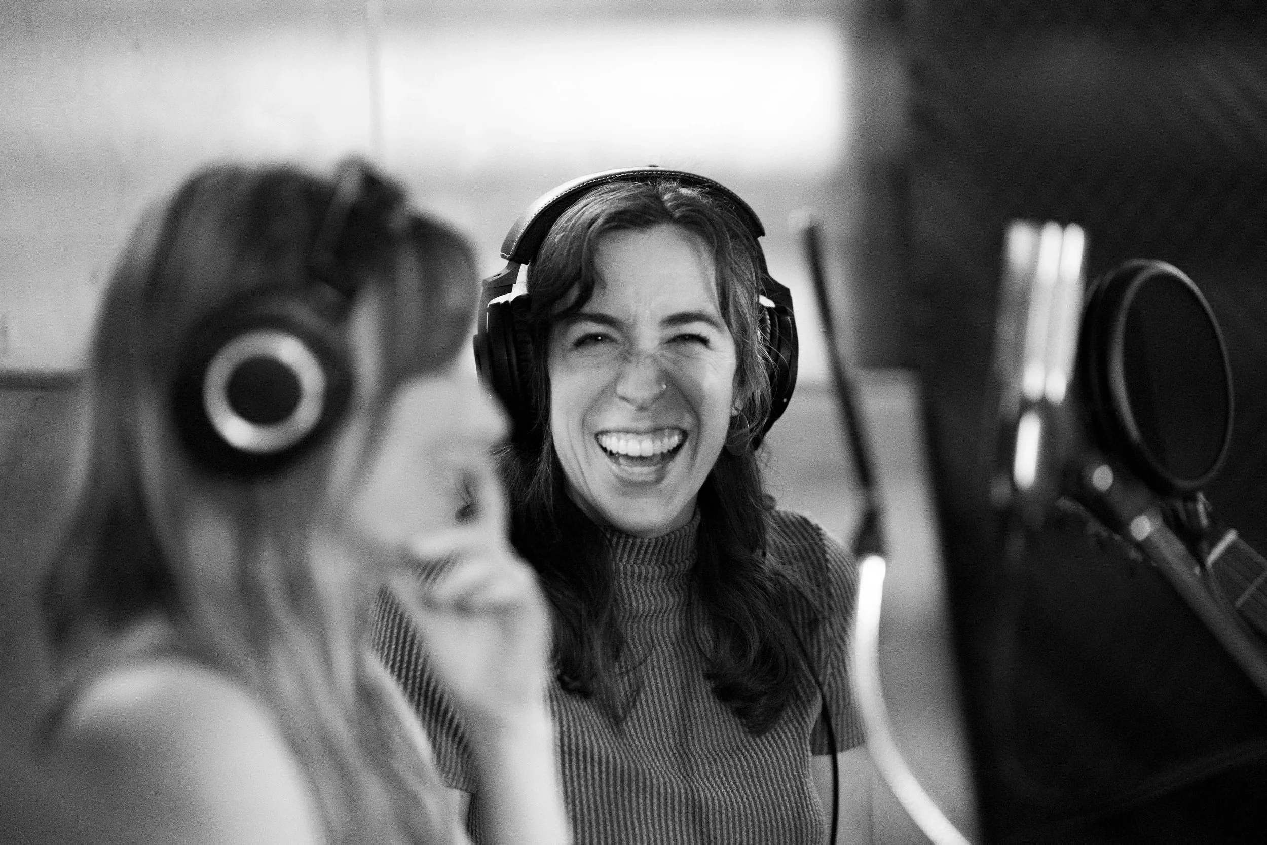 Two women in a recording studio, one woman is laughing and the other woman is adjusting headphones.