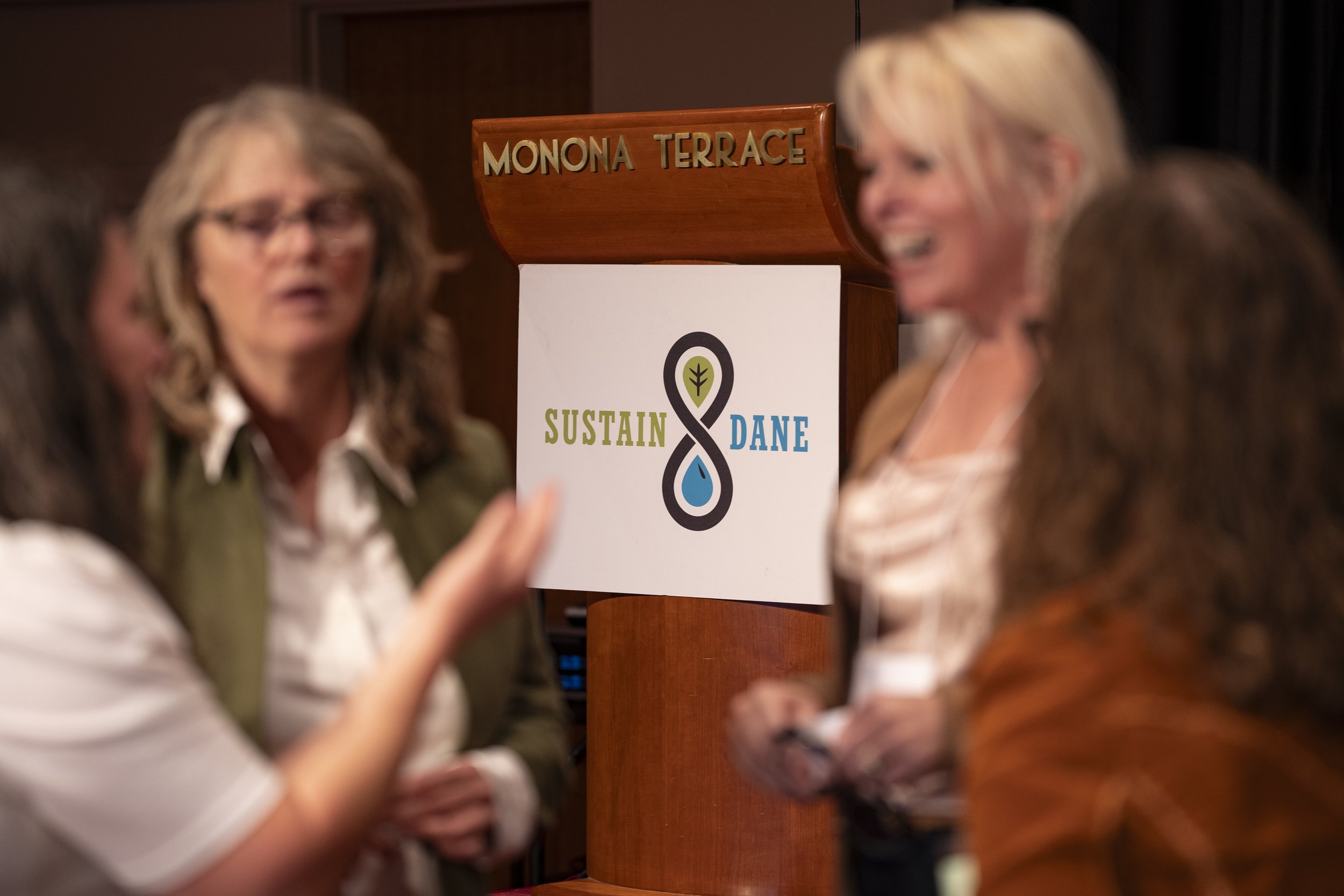 Group of women in conversation near a sign that reads 'MONONA TERRACE' and 'SUSTAIN DANE.'