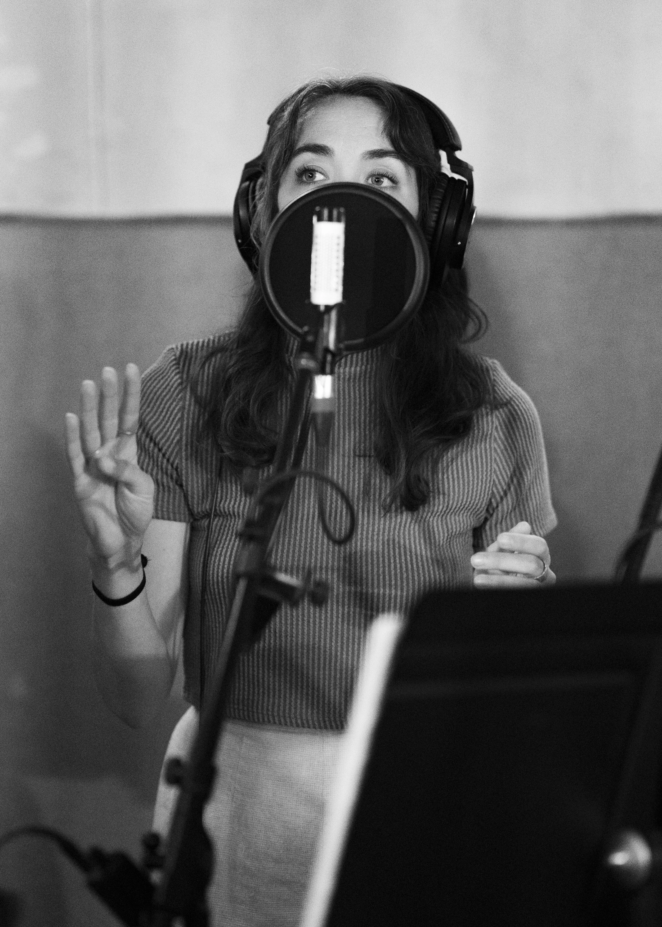 A woman with headphones in a recording studio, standing in front of a microphone with a pop filter, singing or speaking, with her right hand raised.