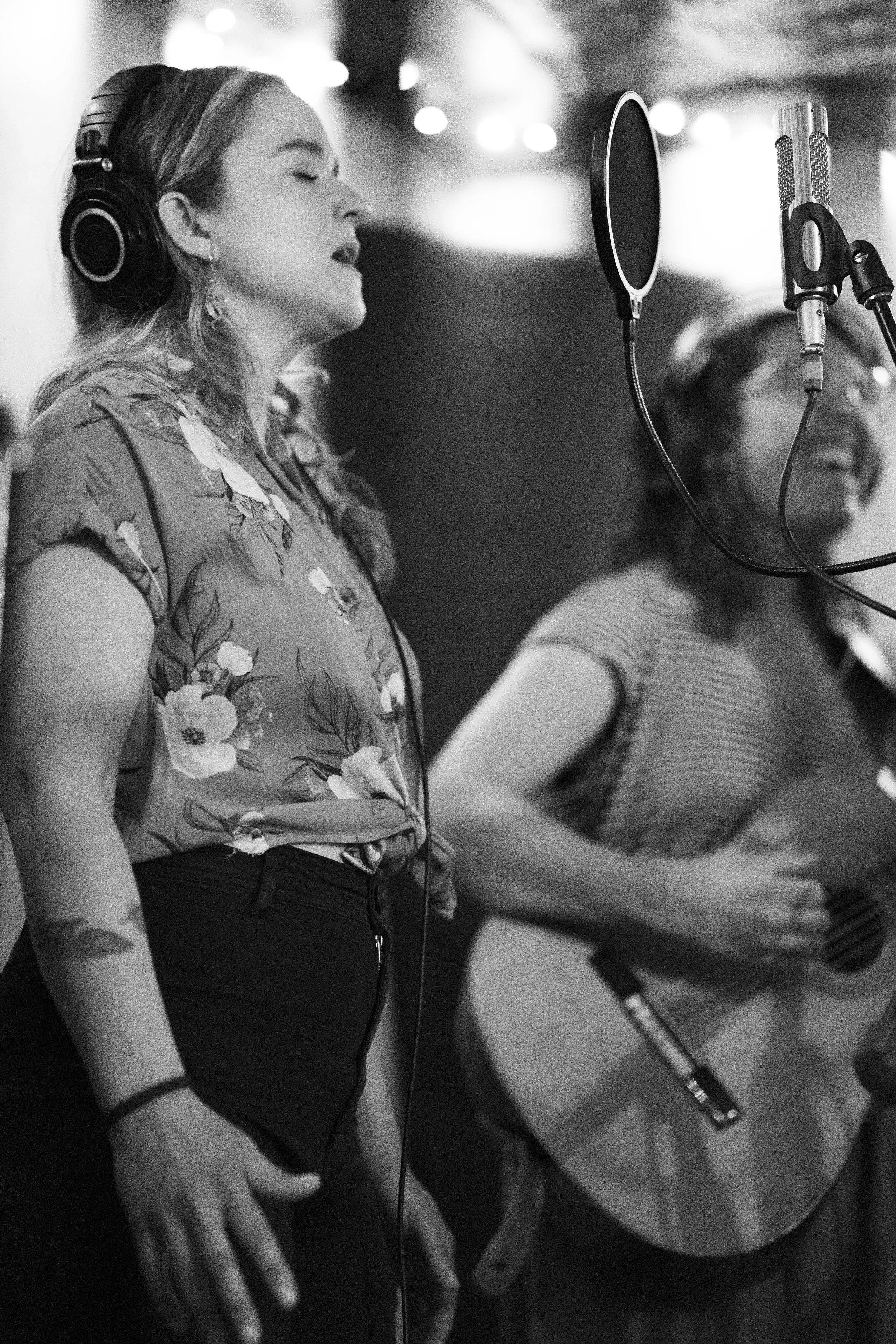 Two women singing into microphones in recording studio, one wearing headphones and floral blouse, the other playing guitar and wearing striped shirt, in black-and-white photo.