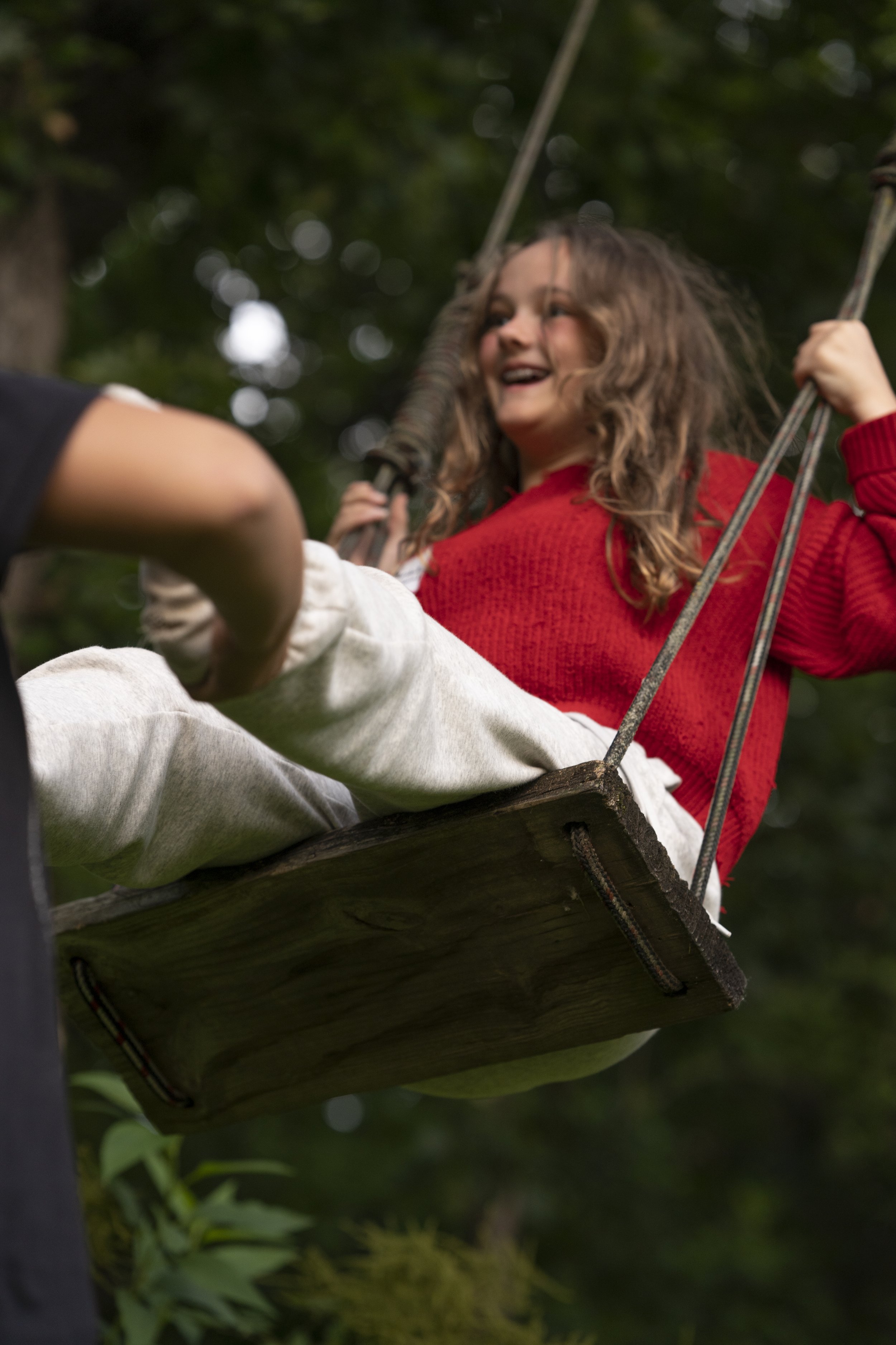 Young girl smiling on a wooden swing hanging from a tree, outdoors in a lush green forest.