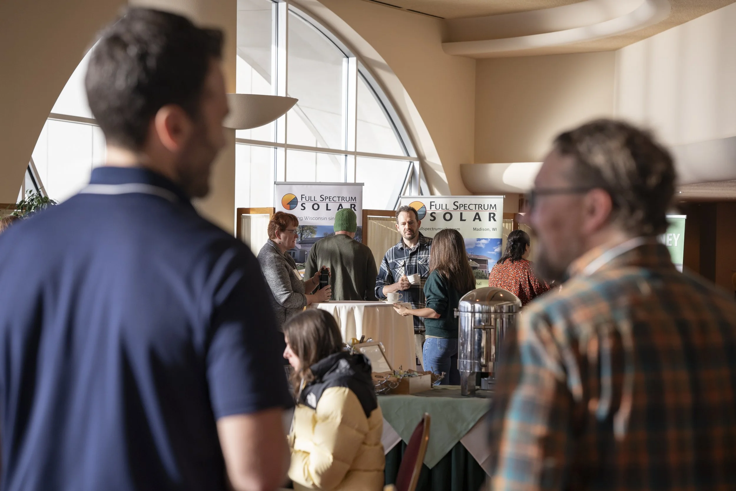People attending an indoor event or conference with booths, including one for Full Spectrum Solar, in a room with curved ceiling and large windows.