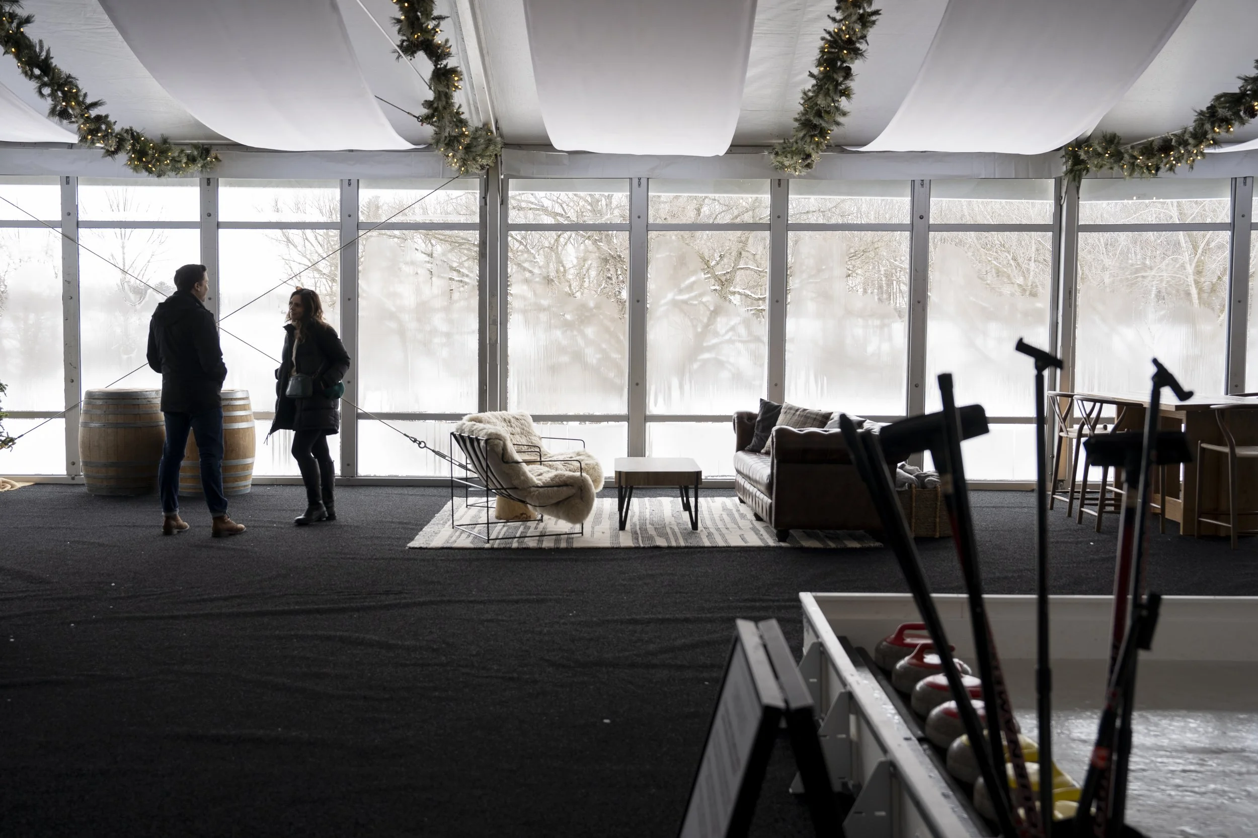 Two people talking inside a large windowed indoor space decorated for winter or holiday season, with snow visible outside. The room has lounge seating and a bar area with golf clubs in the foreground.
