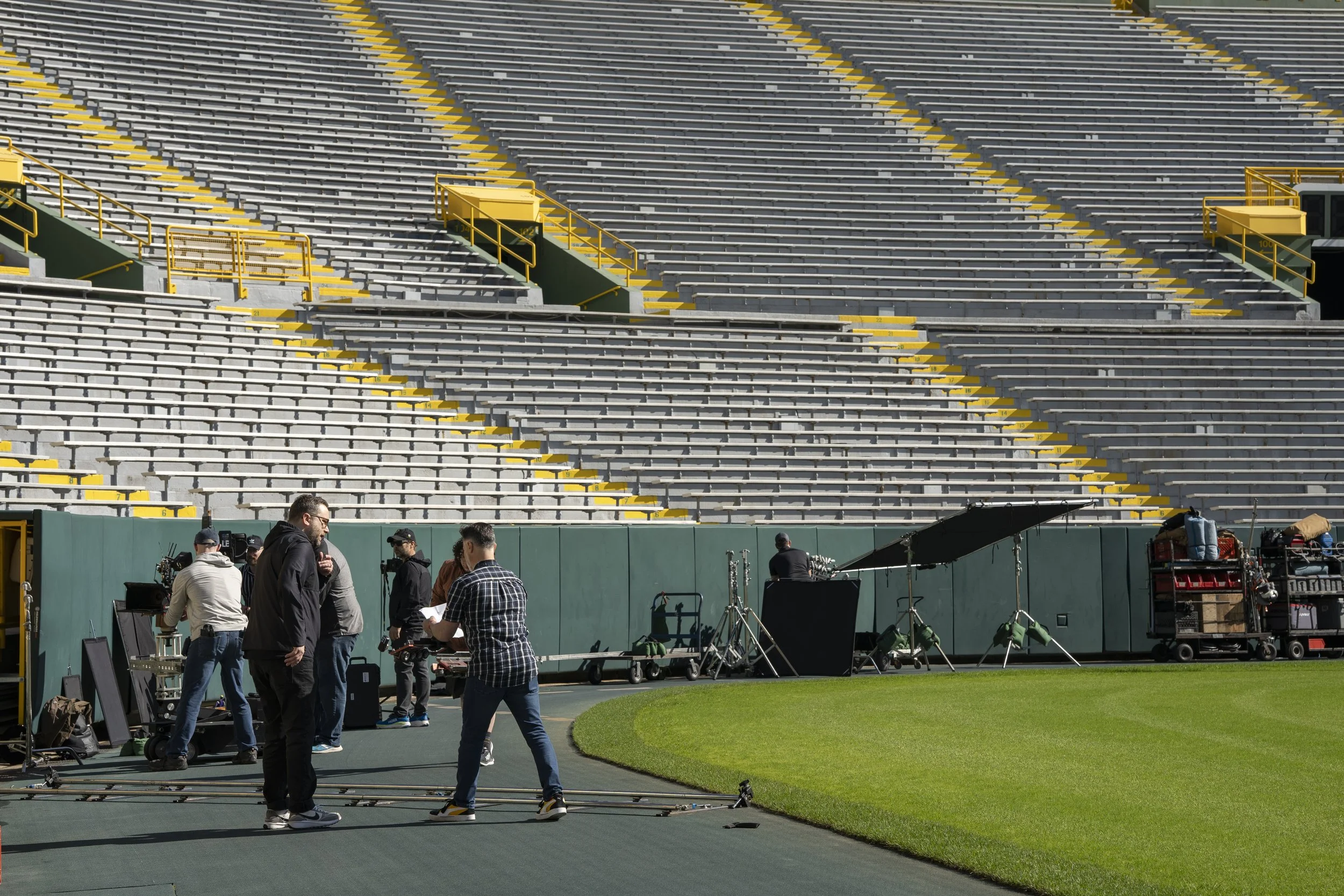 Film crew setting up equipment on a sports field in front of empty stadium seats.