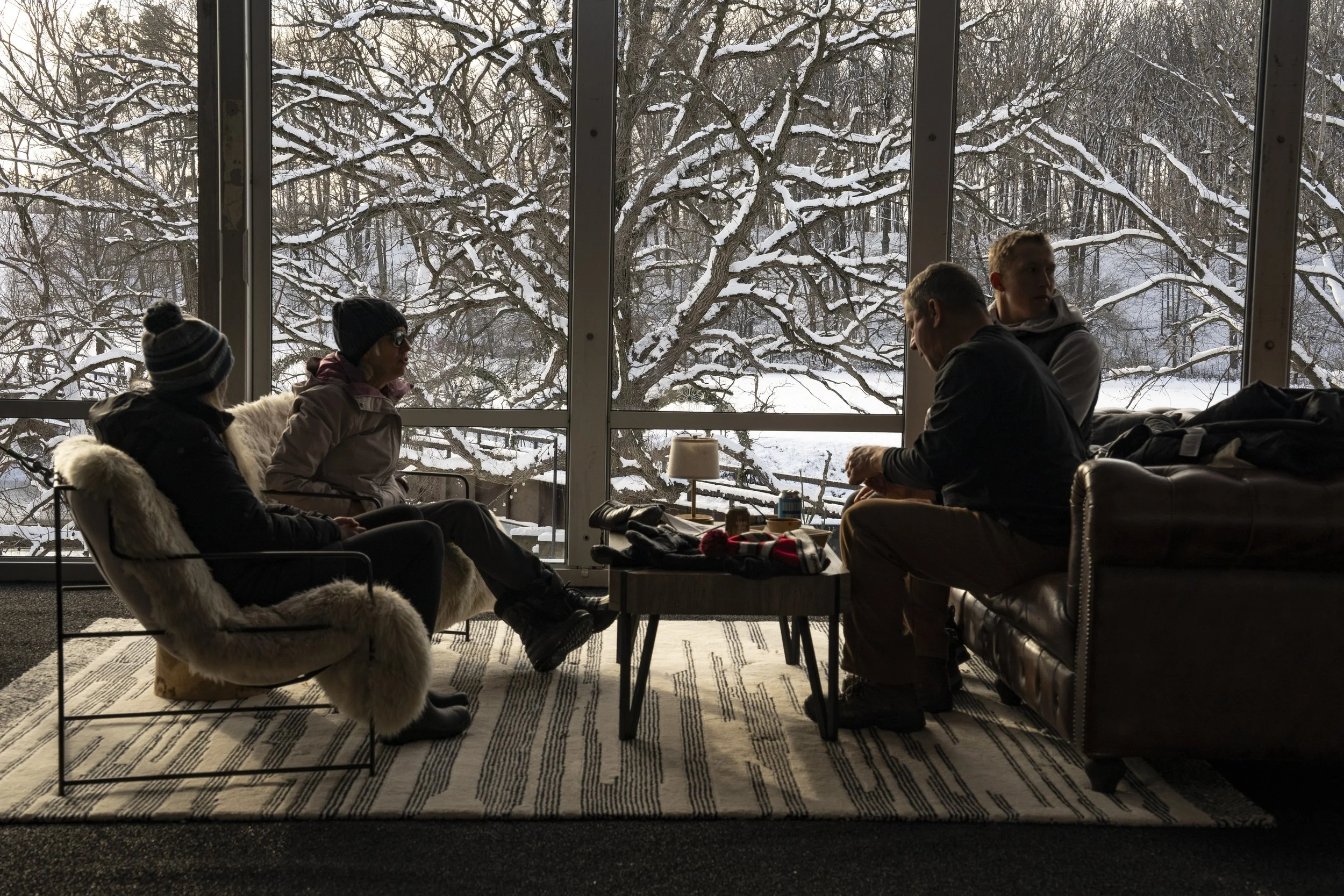 Four people sitting inside a lounge area with large windows showing a snowy landscape and snow-covered trees outside.