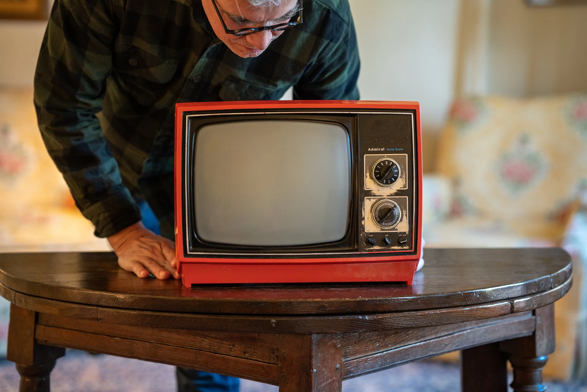 A man in glasses leaning over a small vintage television on a wooden table.
